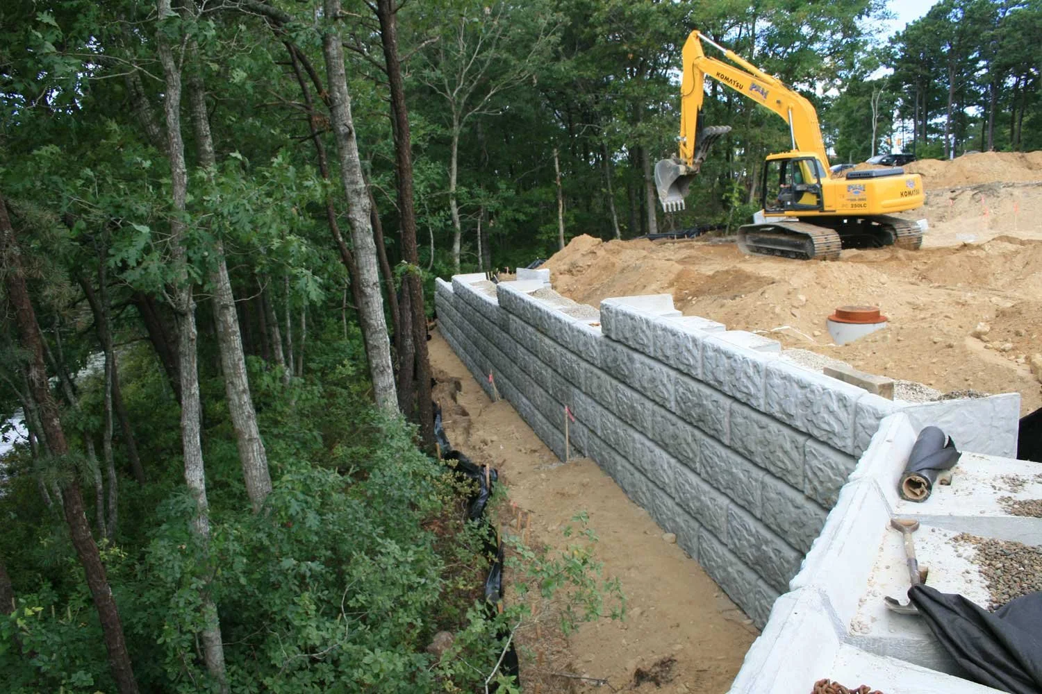 Construction site with a yellow excavator building a retaining wall built by Iron City Retaining Walls, a commercial retaining wall contractor, with large concrete blocks among trees.