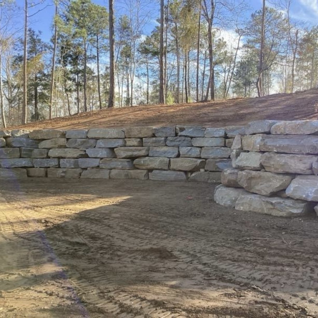 A stone retaining wall built by Iron City Retaining Walls, a commercial retaining wall contractor, in a wooded area with trees and a partly cloudy sky in the background.