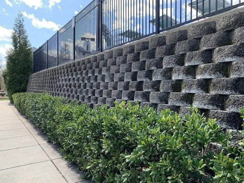 Block retaining wall by Iron City Retaining Walls, a retaining wall contractor in Birmingham, Alabama with a metal fence on top, green bushes along a sidewalk, and a tall tree in the background.