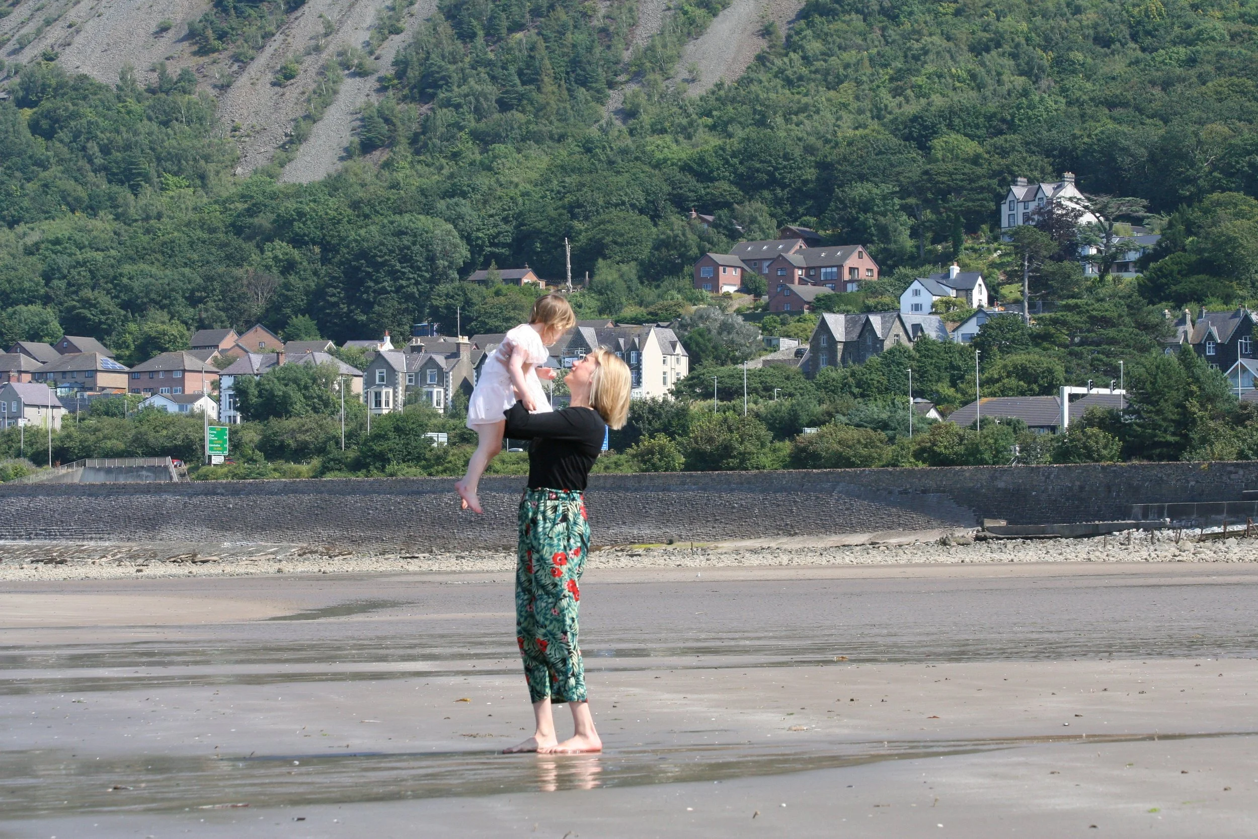 A woman lifting a toddler on a beach, with houses and wooded hills behind