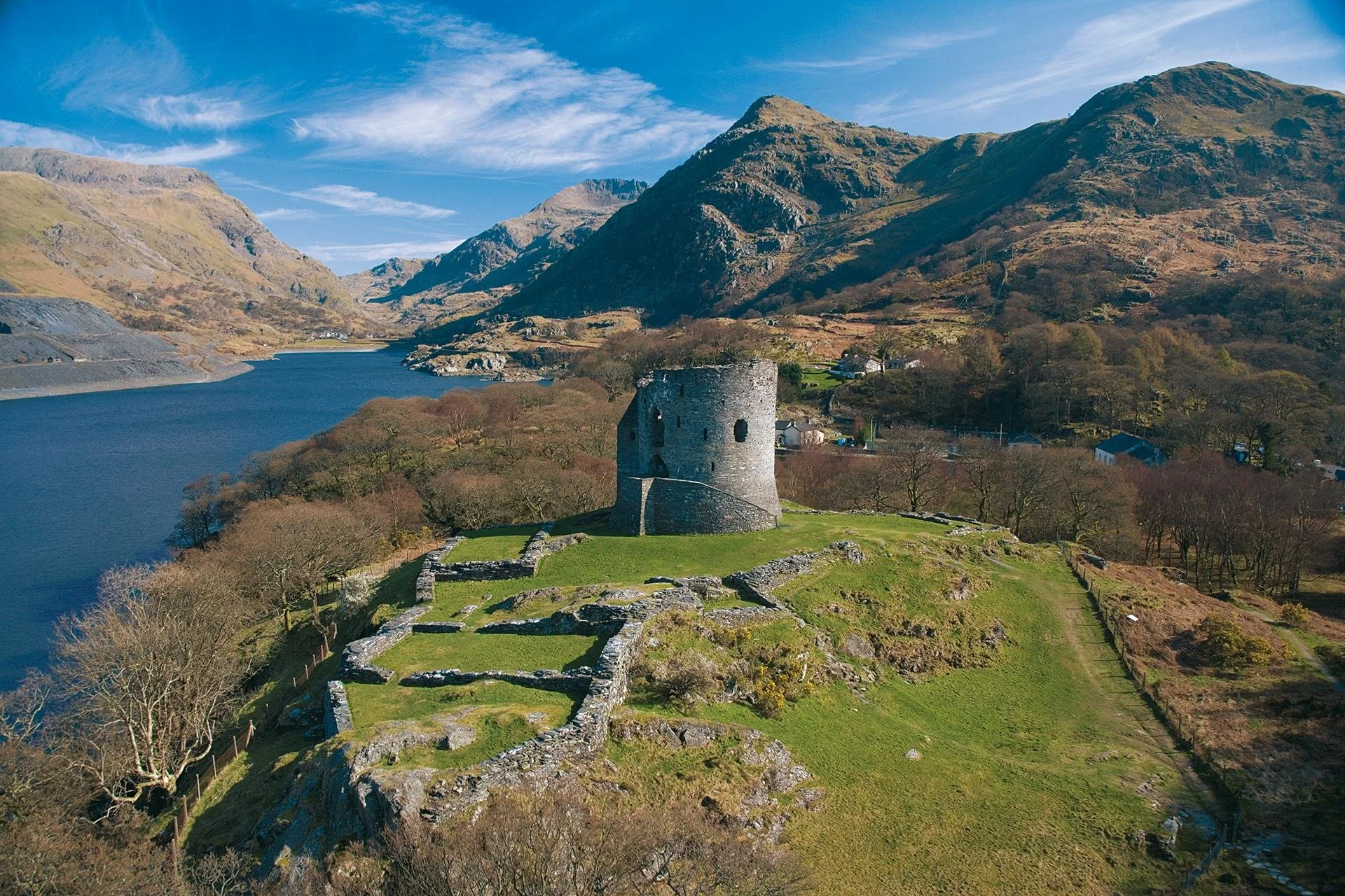 An aerial view of the round tower of Dolbadarn Castle on a grassy hill beside a lake, with mountains behind