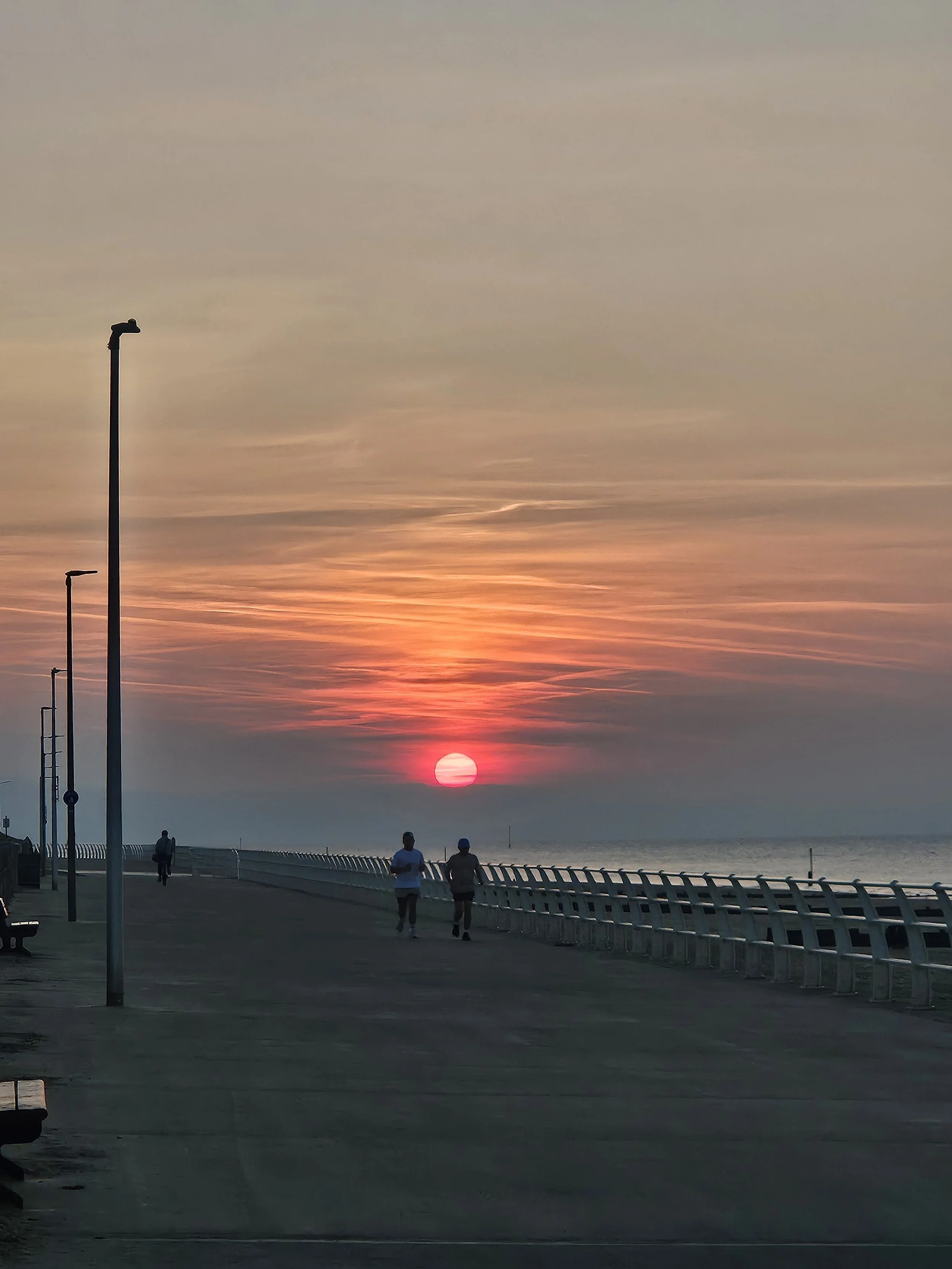 Sunset over the sea beyond a promenade, with two people walking along it