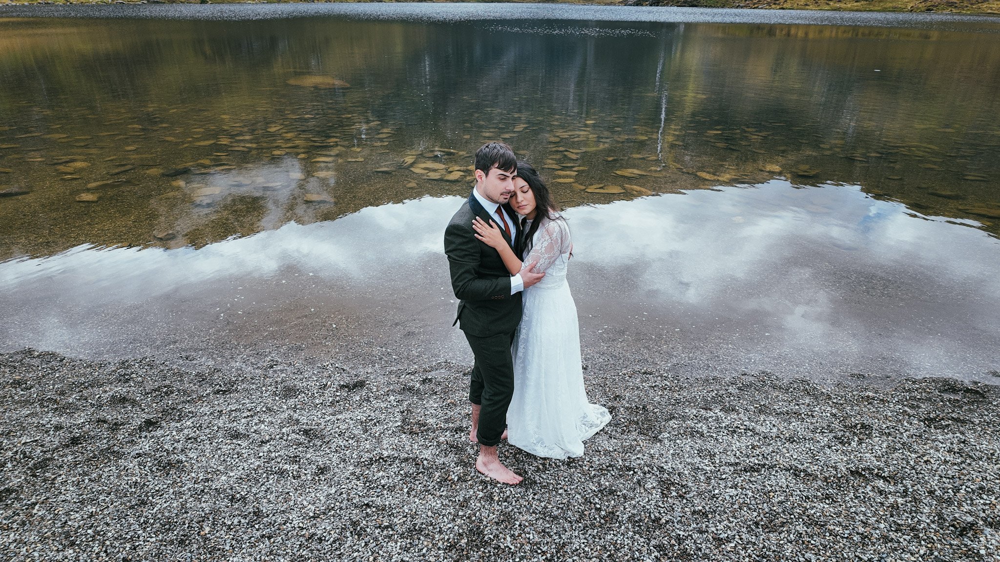 A couple embracing on a pebbly lakeshore with clouds reflected in the water