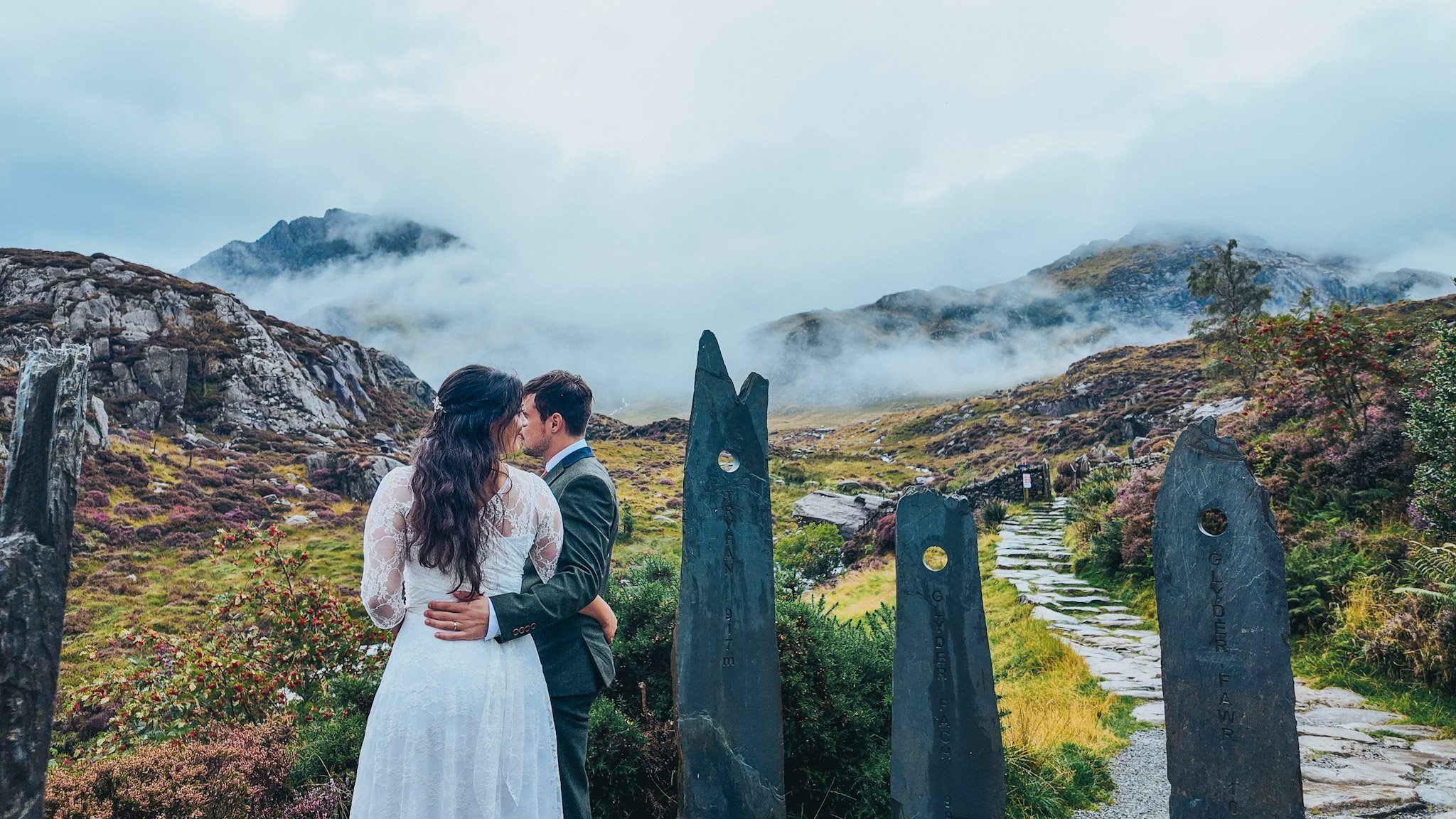 A couple embracing on a stone path between standing stones, with mist over the mountains