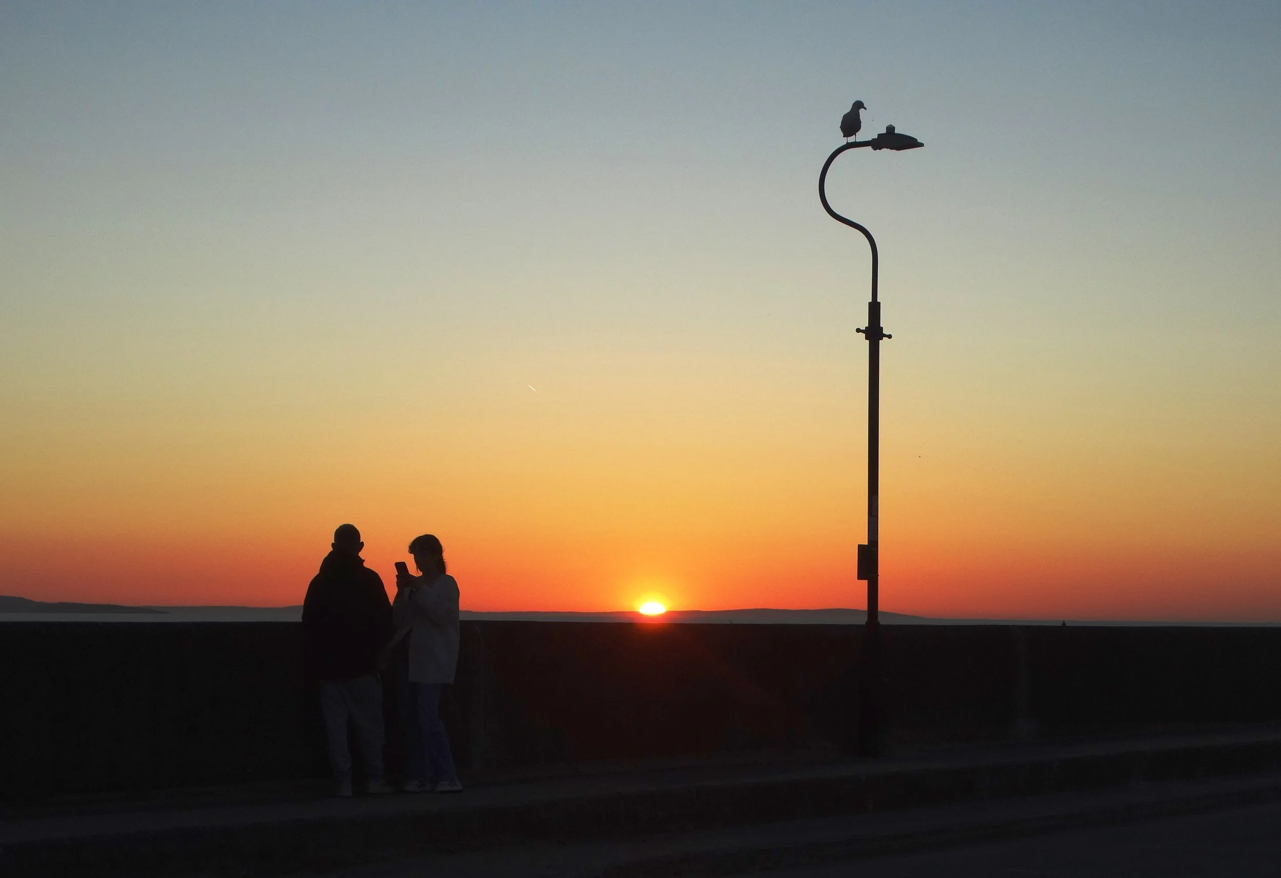 Two people silhouetted against an orange sunset beside a lamp post at West Bay, Llandudno
