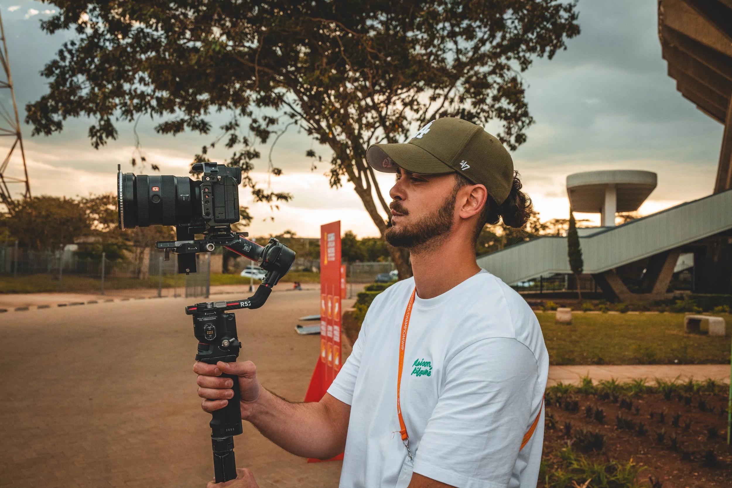 Un homme tient une caméra stabilisée lors d'une prise de vue en extérieur, avec un ciel nuageux et un arbre en arrière-plan.