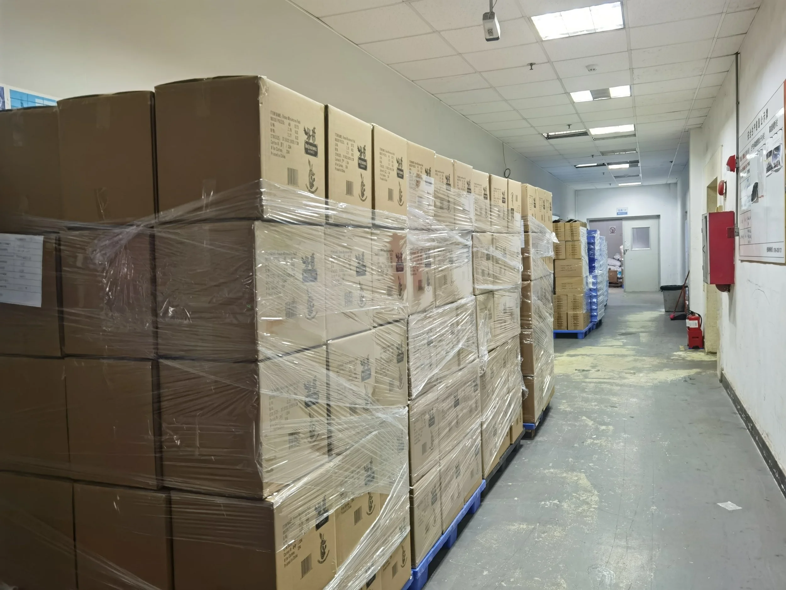 A corridor with stacked cardboard boxes wrapped in plastic on blue pallets, a mop and bucket at the end, and various signs and a fire alarm on the wall.