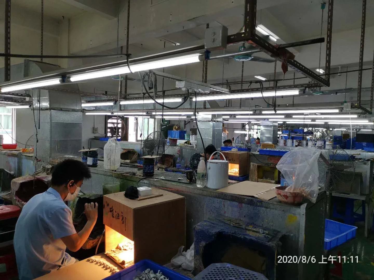 Indoor workshop or factory with workers, some wearing masks, working at workbenches with tools and materials, fluorescent lighting, and industrial equipment.