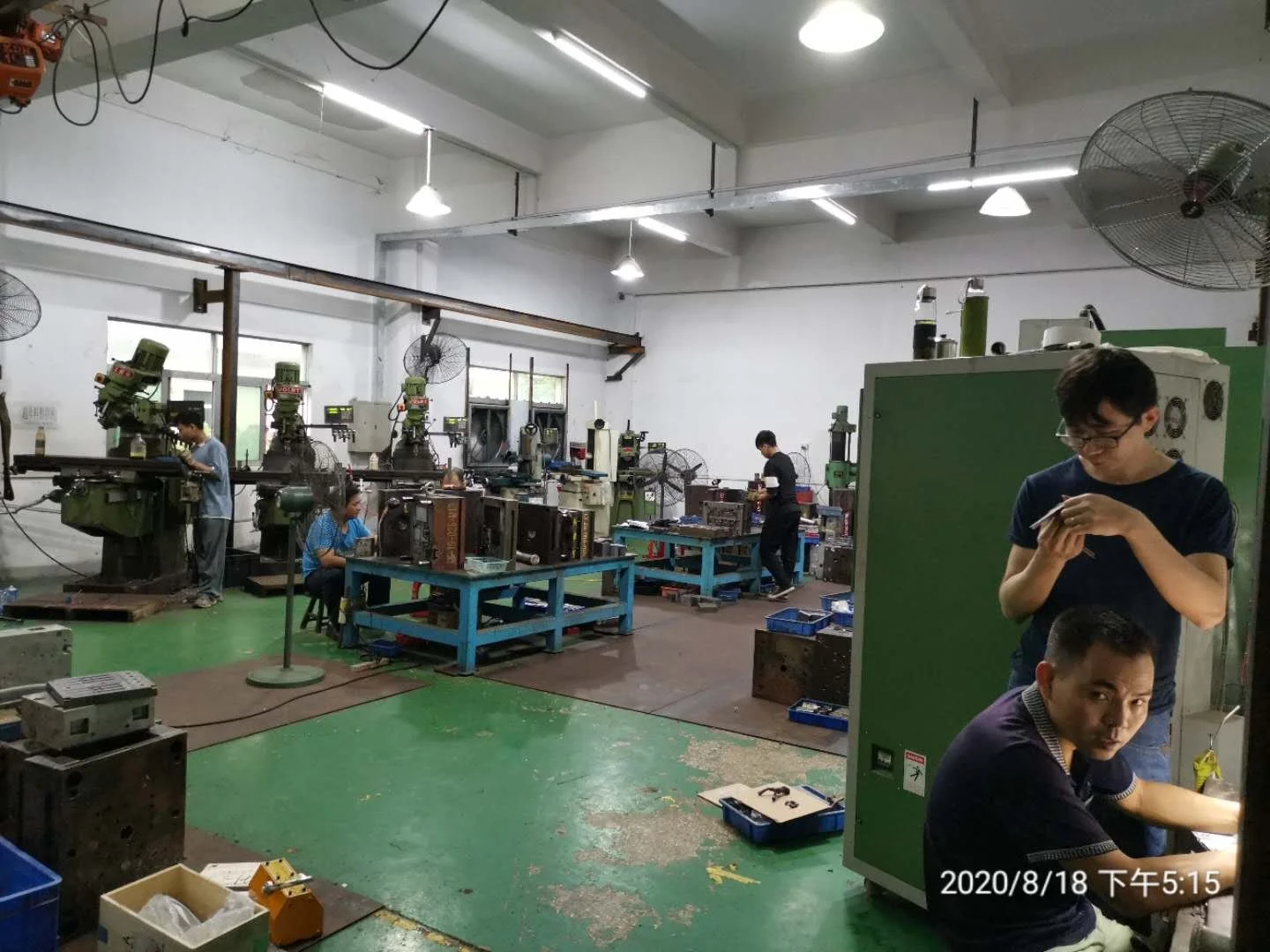 View of a mechanical workshop with several workers operating large machinery and tools, some sitting and some standing, in a well-lit space with white walls and fluorescent lighting, with various boxes and equipment on tables and the floor.