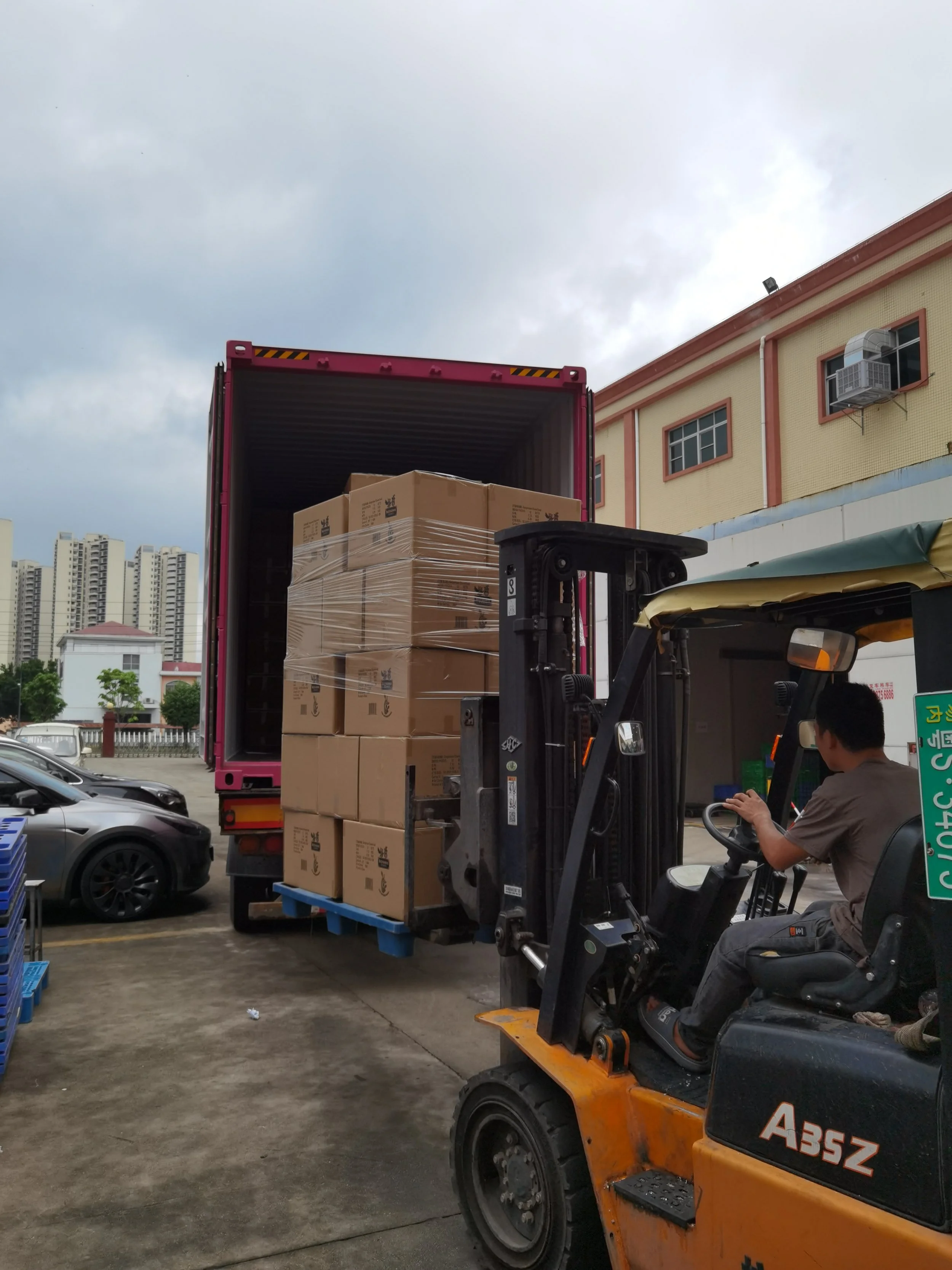A forklift operator is moving boxes from a truck onto the ground at a loading dock. The truck's back doors are open, revealing stacked boxes wrapped in plastic. The scene takes place outdoors in a parking lot near a building with an air conditioning 