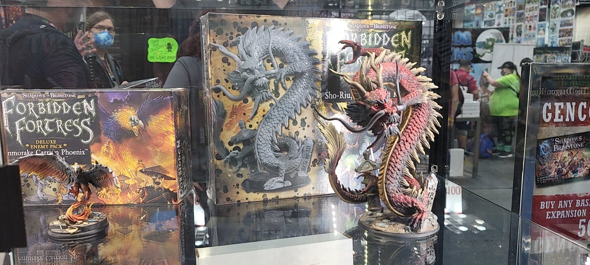 Display of miniature models and game boxes of Forbidden Fortress, featuring detailed dragon and phoenix figurines, at a convention or store with people in the background
