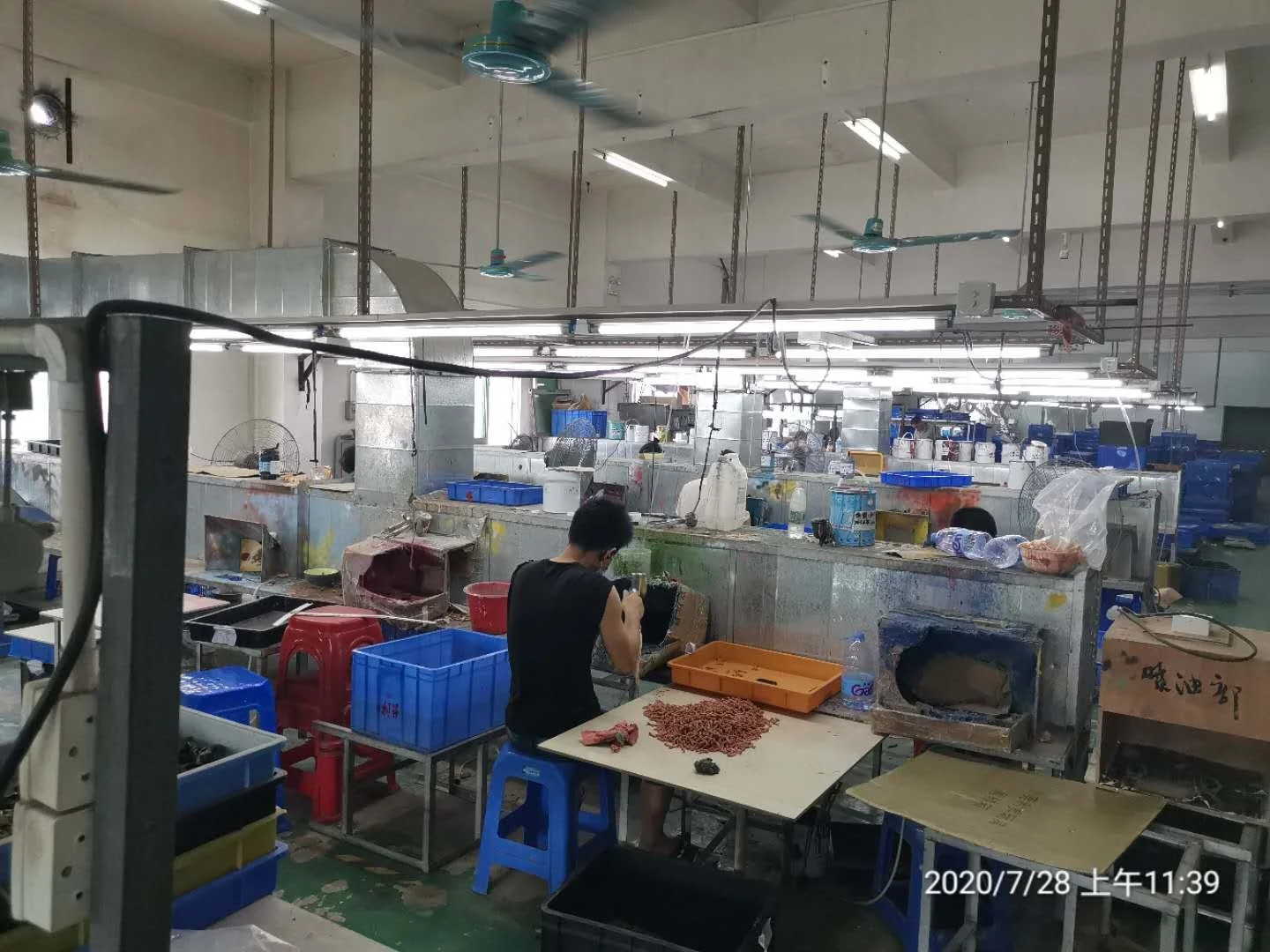 An industrial workspace with a person working at a sink or workstation, surrounded by containers and equipment, with ceiling fans and bright fluorescent lights overhead.