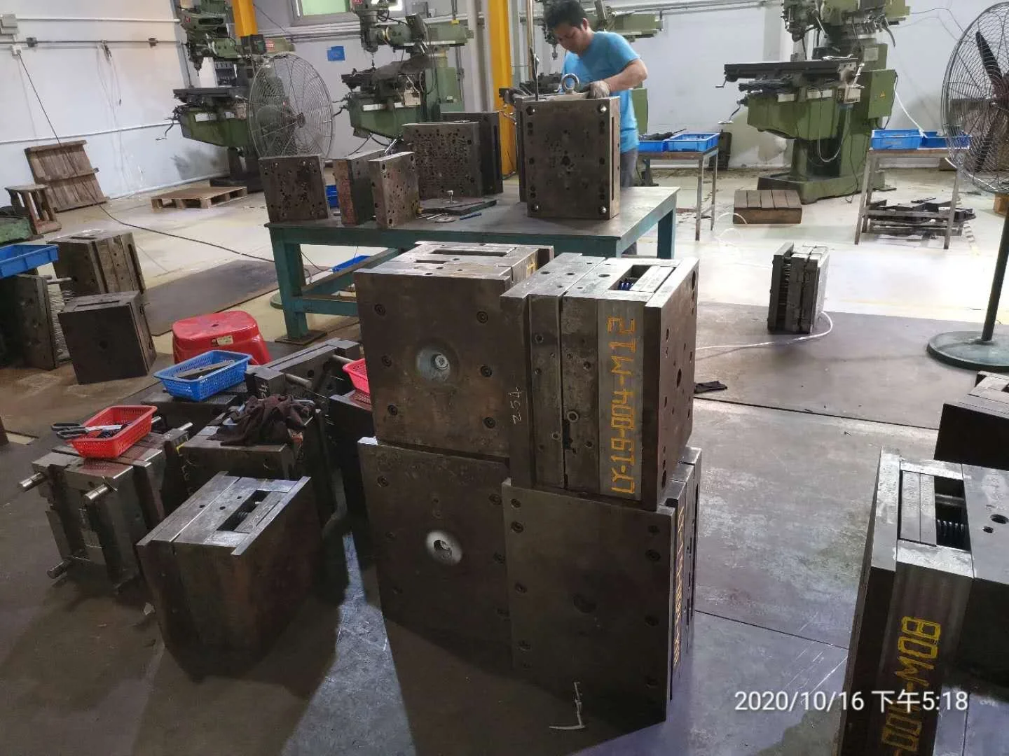 A metalworking factory with large molds, tools, and equipment. A worker in a blue shirt is working in the background, surrounded by industrial machinery and fans.