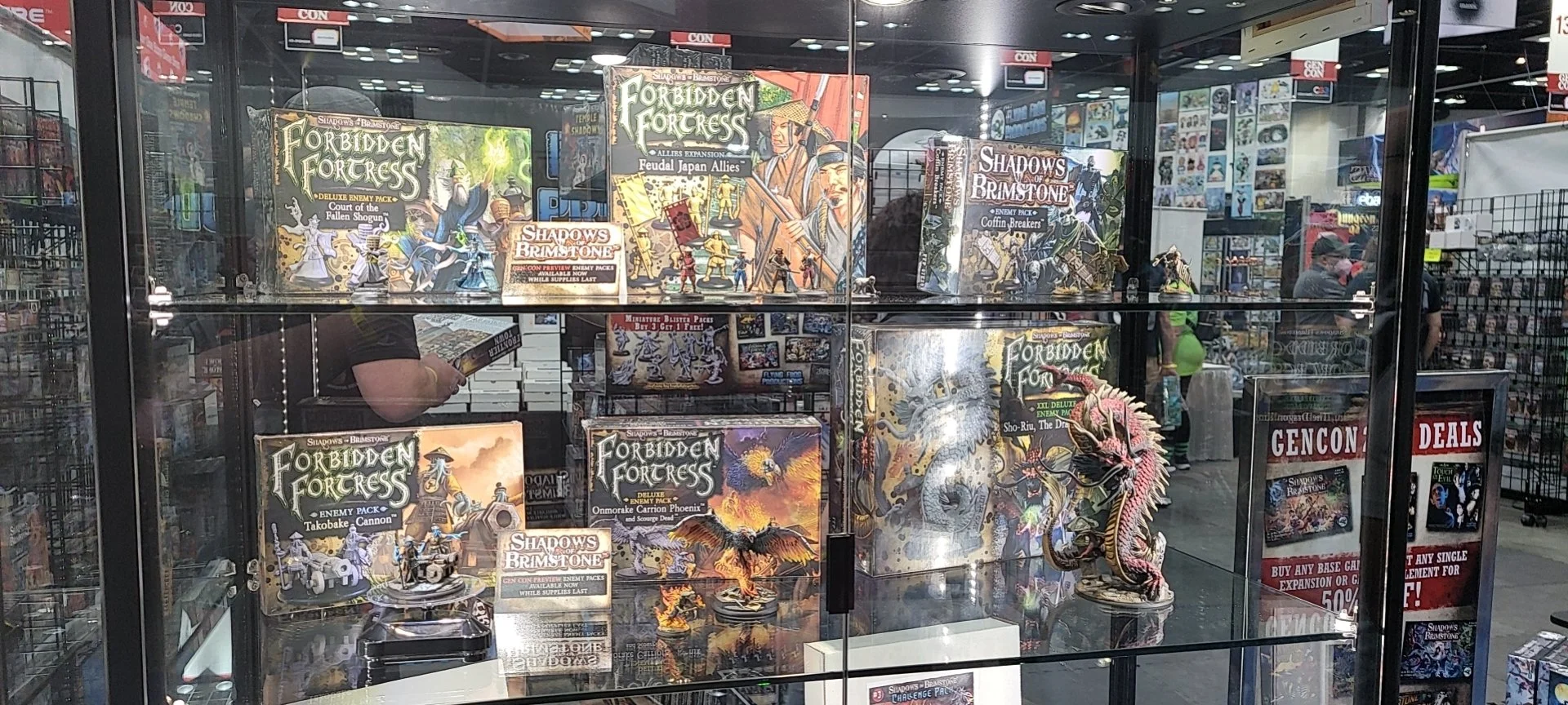 A glass display case filled with boxed sets and miniatures of the board game 'Shadows of Brimstone,' with promotional signs and a dragon miniature inside the case.