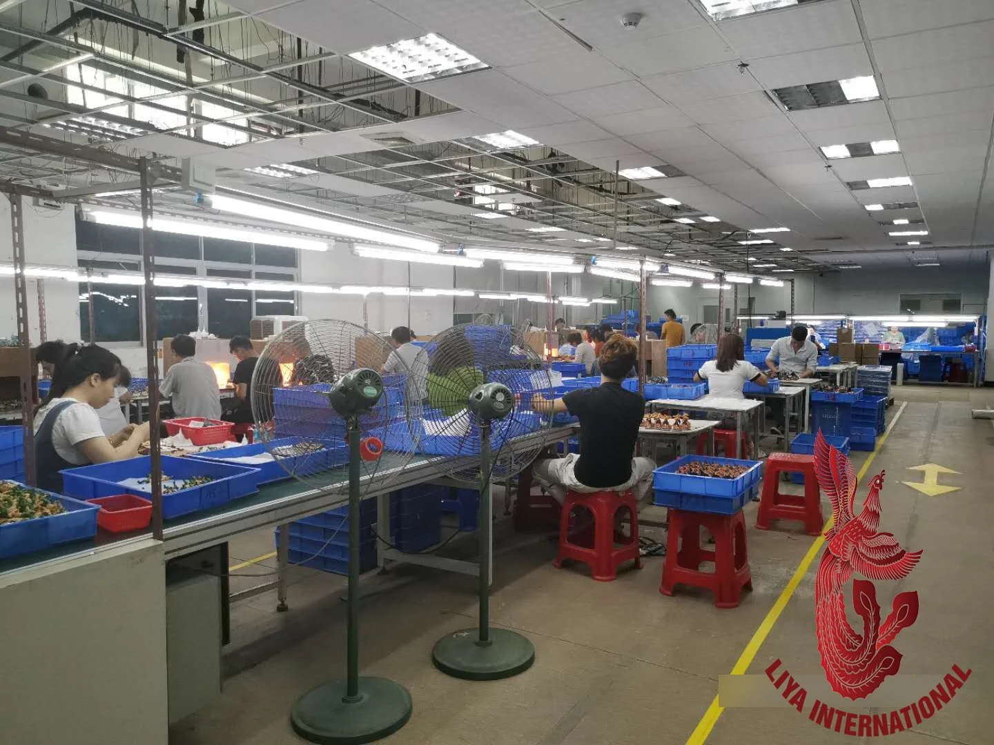 People working at assembly lines in a factory, with blue plastic bins, fans, and bright overhead lighting.