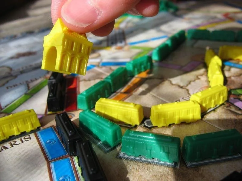 Close-up of a hand holding a yellow train piece over a game of Monopoly with various colored train pieces on the board.