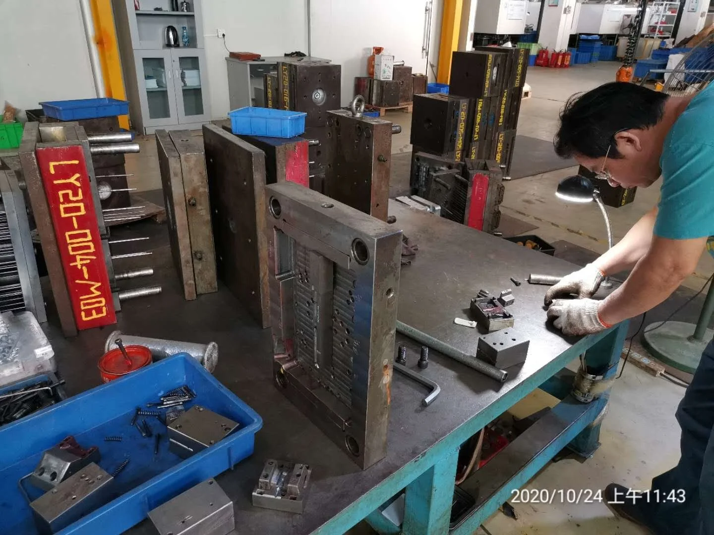 A person working at a metal workbench in a workshop, assembling or repairing metal components surrounded by various metal parts and tools, with shelves and storage in the background.