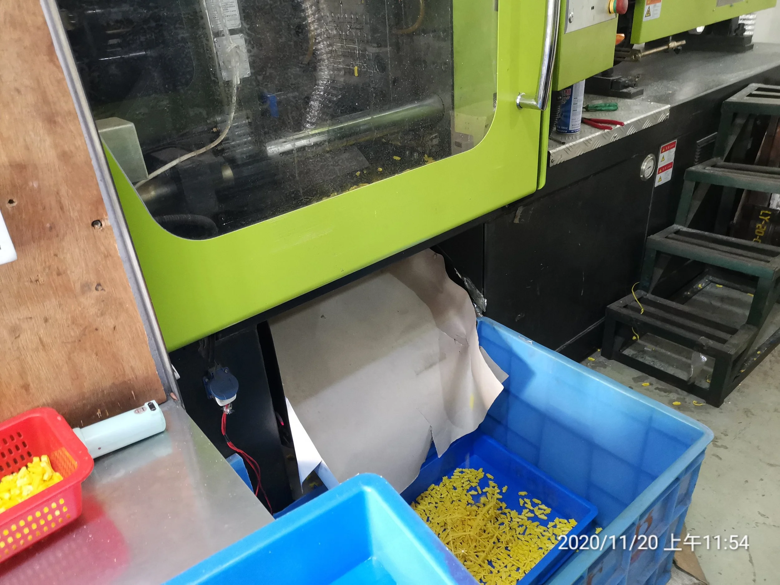 A manufacturing workspace with a machine enclosed in green and black casing, plastic yellow pieces in blue containers, a step ladder, and scattered small yellow parts on the floor. The time and date are displayed as 2020/11/20 11:54.