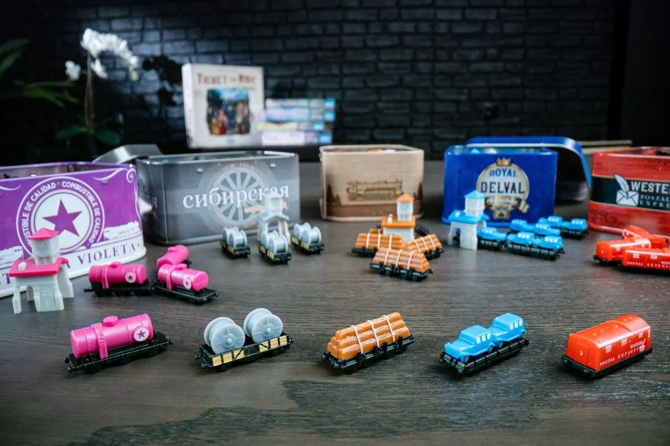 Collection of small toy train cars in various colors including purple, gray, orange, blue, and red, arranged on a dark wood surface with metal and cardboard containers in the background.