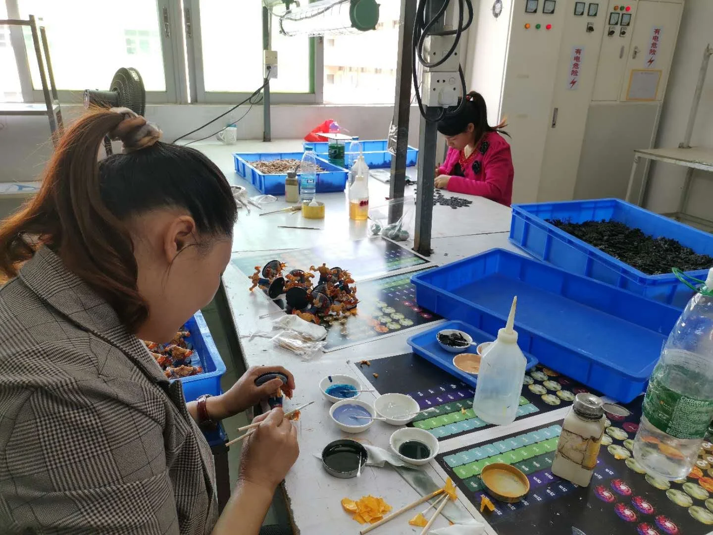 Two women working on crafts at a worktable with various supplies, blue storage bins, and small figurines.