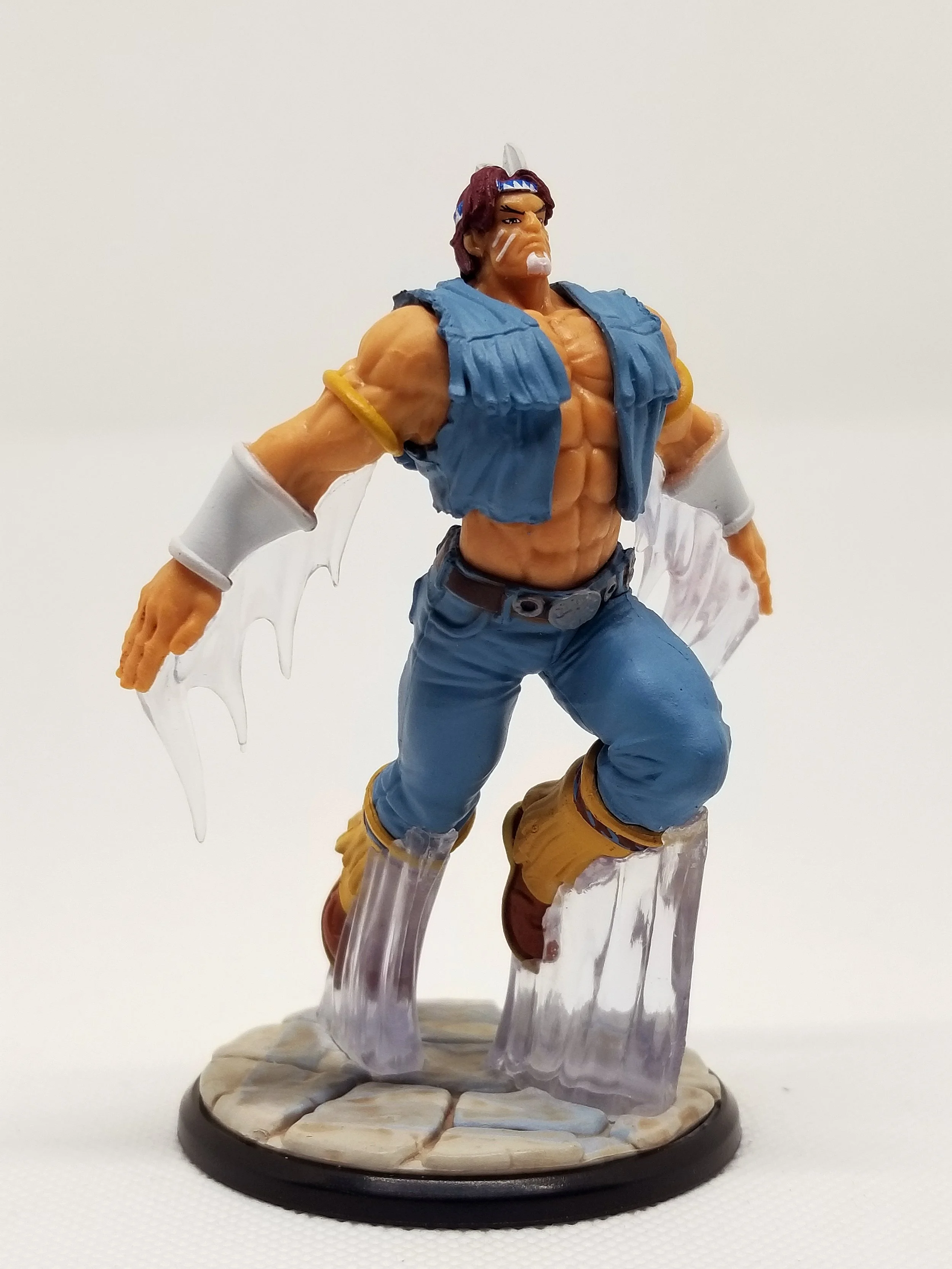 Action figure of a muscular man with a headband, blue vest, blue pants, and boots, standing on a stone-patterned base.