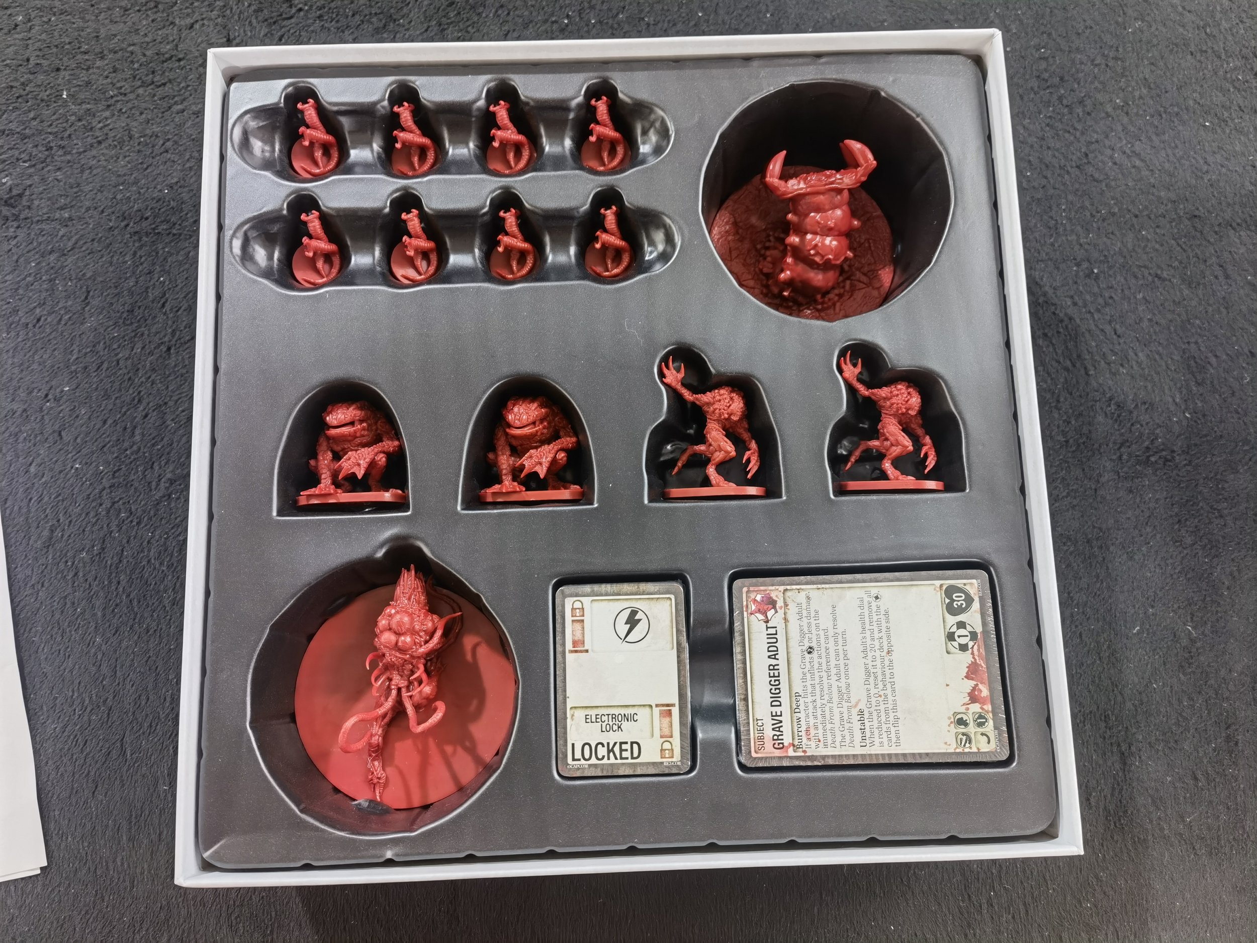 A tabletop game set featuring red miniatures, including monsters and soldiers, arranged in a black plastic tray with designated compartments. The set includes a card with game information and an electronic lock labeled 'LOCKED'.