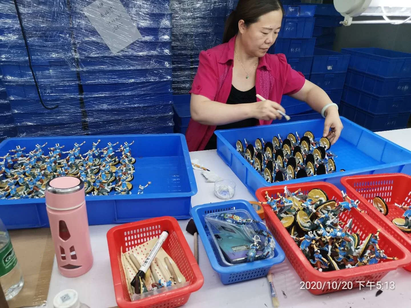 A woman in a pink shirt working with small figurines that appear to be soldiers or warriors, placed in blue and red plastic baskets on a table. The woman is handling or assembling the figurines, which are detailed with colorful uniforms and weapons. There are containers, a water bottle, writing tools, and other items on the table.