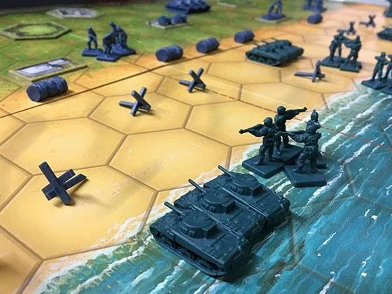 A strategic tabletop war game with hexagonal tiles depicting land and water, military figures, tanks, and other game pieces.