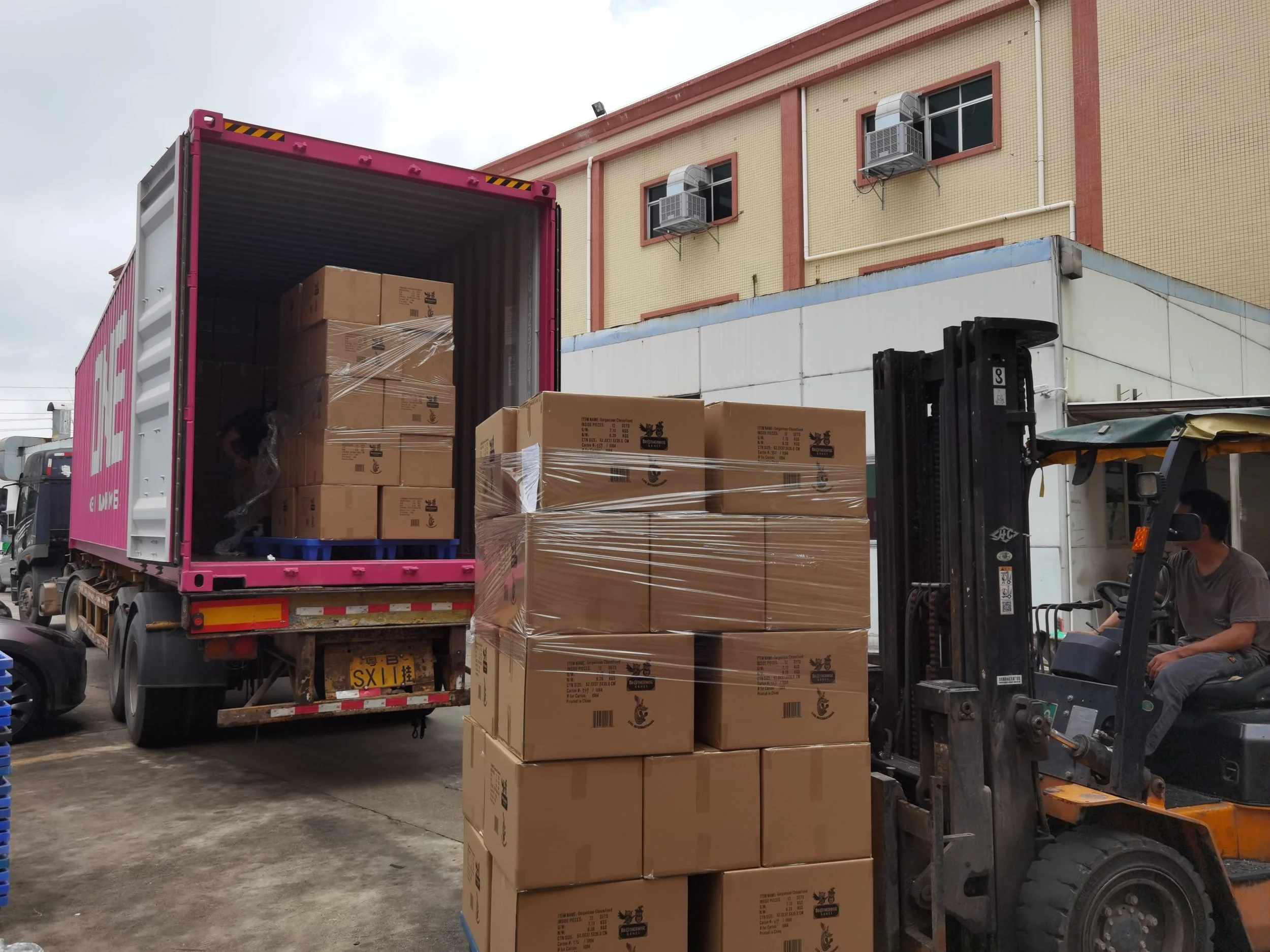 A worker driving a forklift moves stacked cardboard boxes next to a pink delivery truck that is being loaded with more boxes for transport at an industrial or warehouse area.