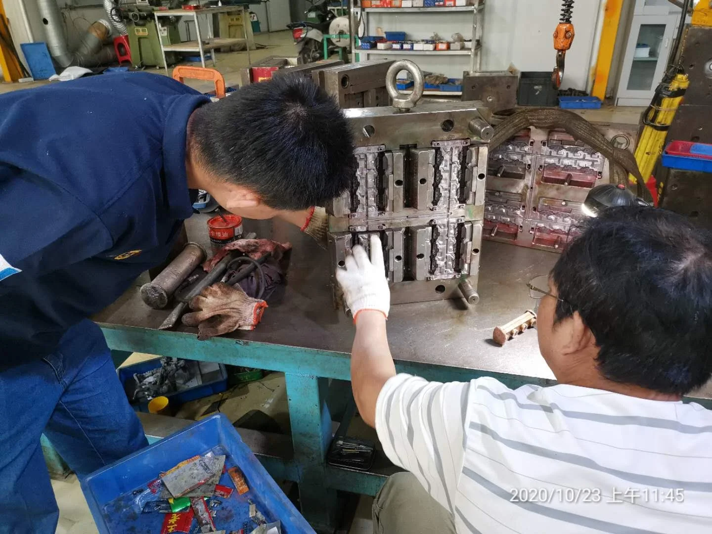 Two men working together to repair a large industrial machine in a workshop. The man on the left is leaning over, and the man on the right is pointing at the machine. The workshop contains various tools, equipment, and shelves in the background.