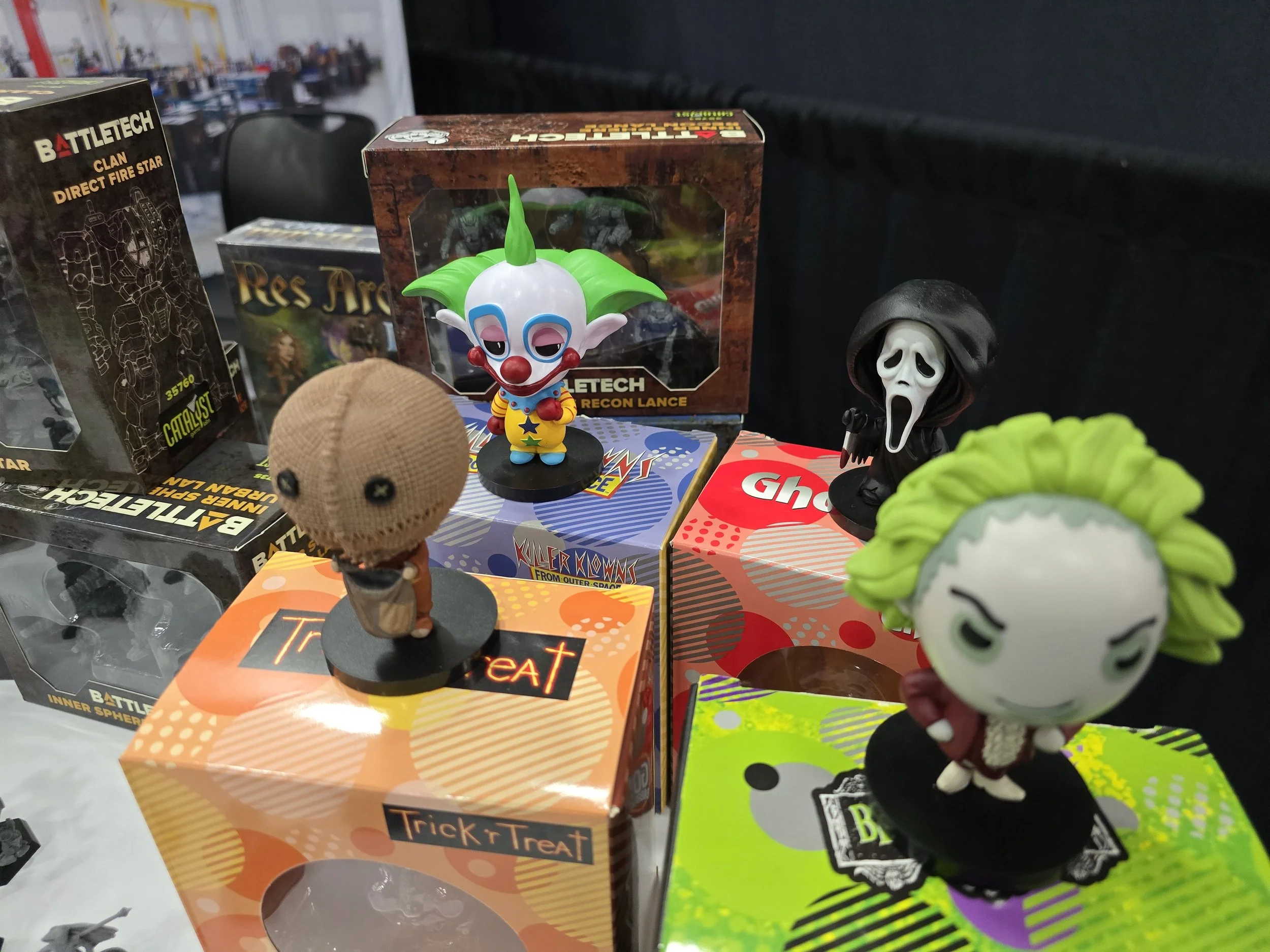 Collection of Halloween-themed figurines and toy boxes, including a clown, a ghost face, a green-haired character, and a doll, displayed on tables with boxes and packaging in the background.