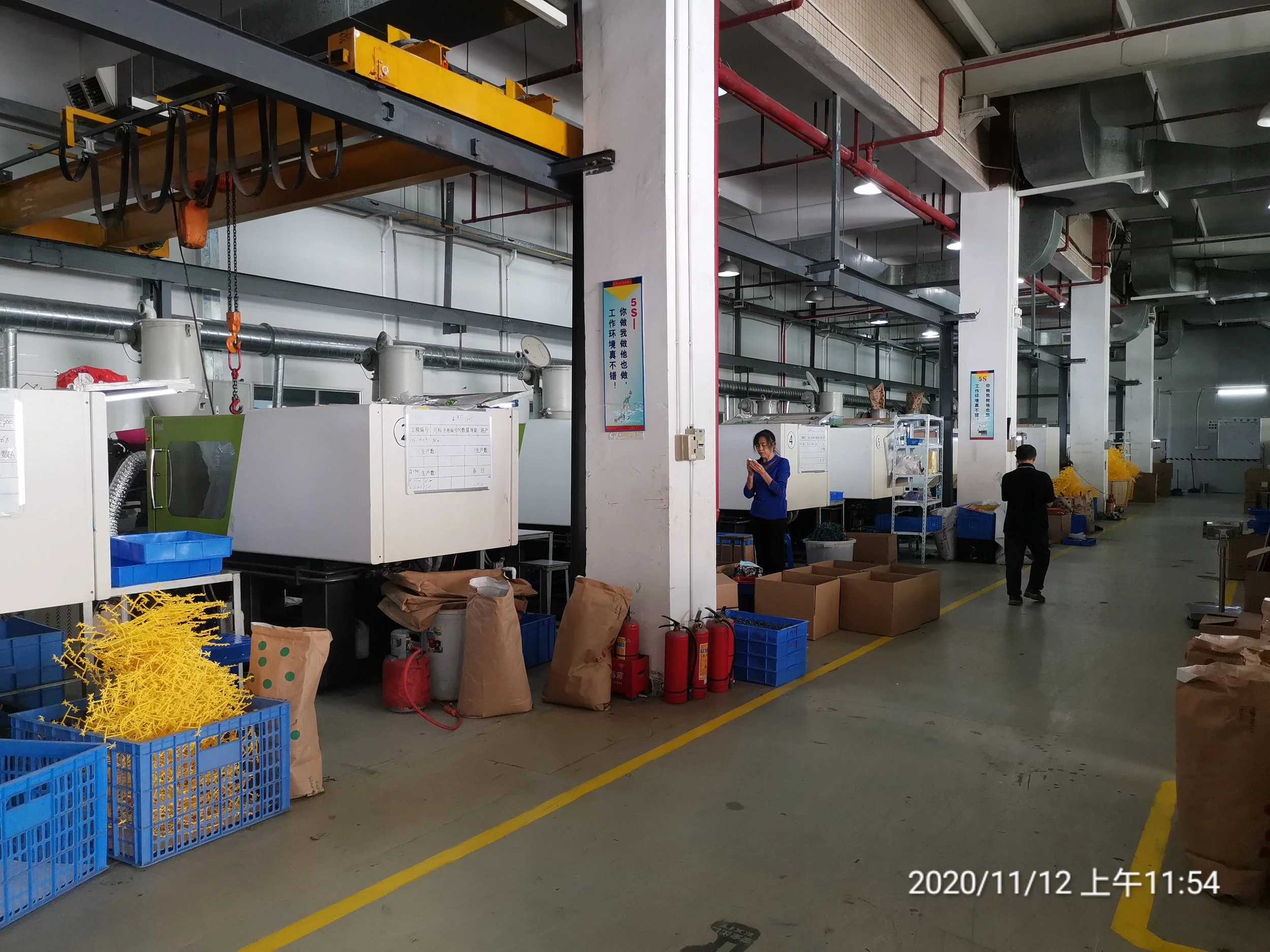 Industrial factory setting with machines, carts filled with yellow plastic sticks, brown paper bags, fire extinguishers, and two workers, one woman and one man, inspecting or working. The space is well-lit with pipes and ventilation systems overhead.