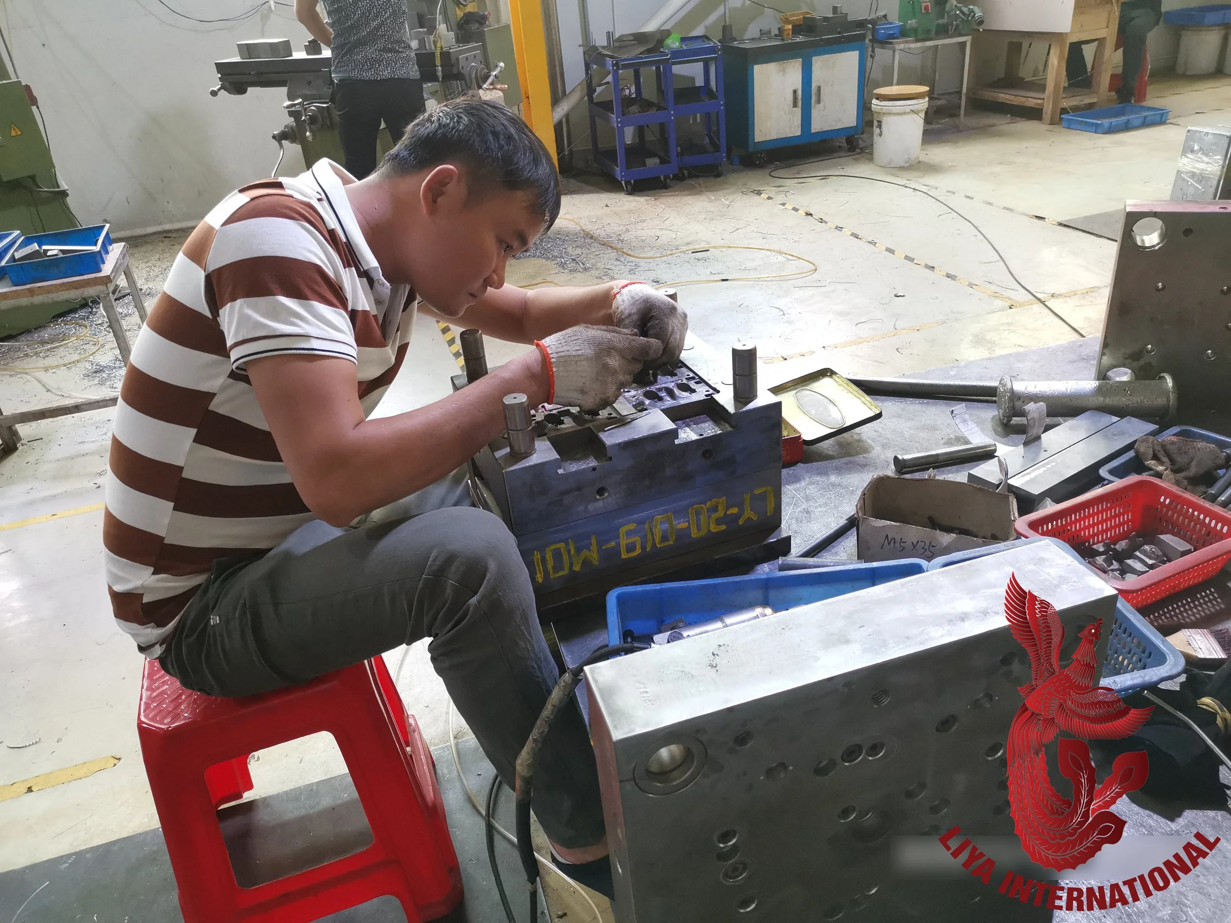 A man in a striped shirt working on a mechanical part at a workshop, seated on a red plastic stool, with various tools and equipment around him.
