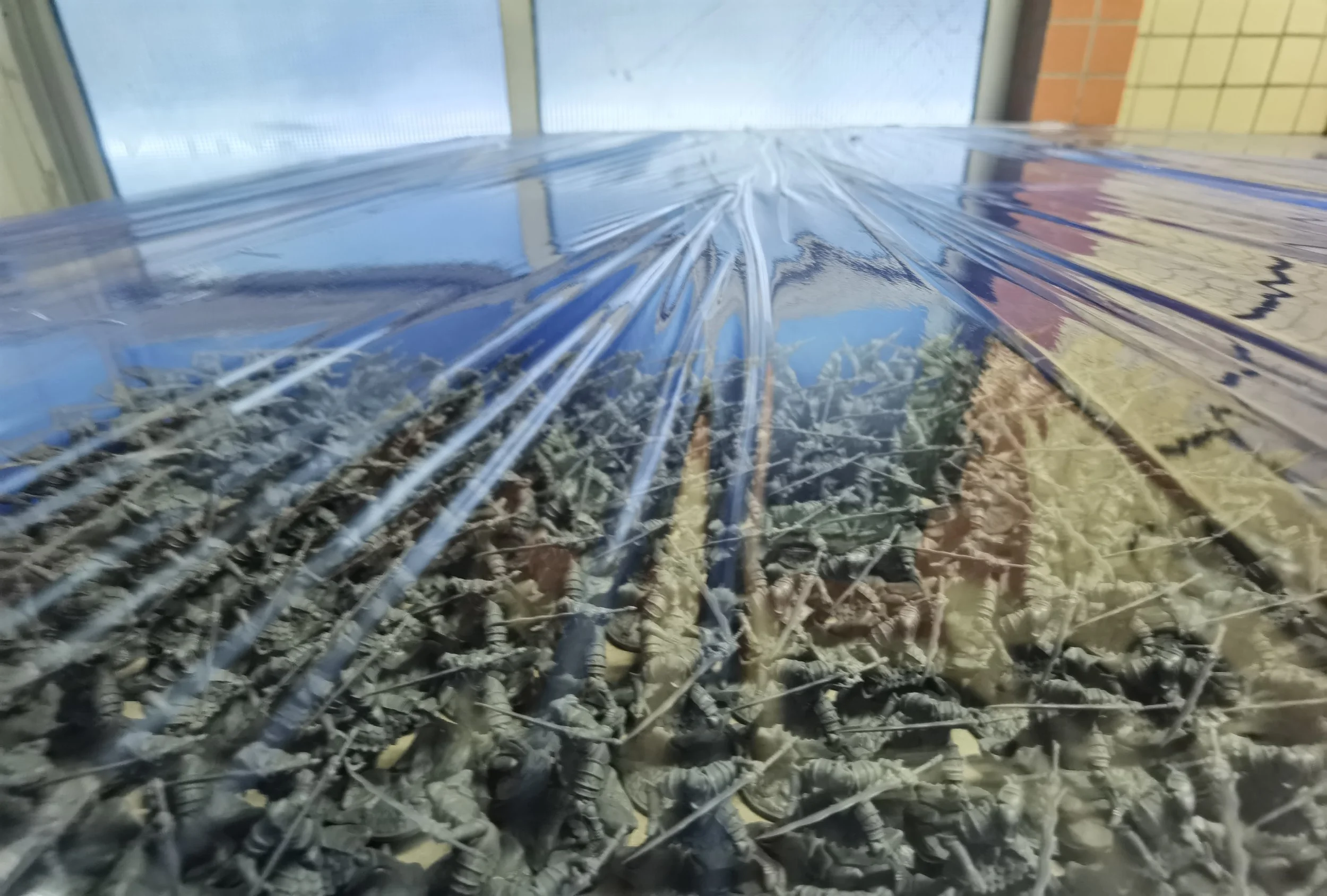 A large number of toy soldiers covered with plastic wrap are visible, with a glass window and brick wall in the background.