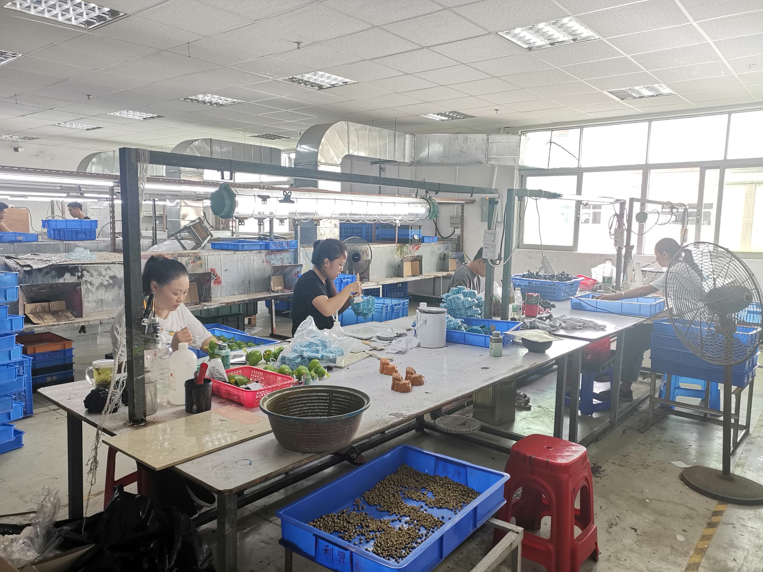 Workers in a factory assembling or packing products at work tables with blue plastic bins and various items.