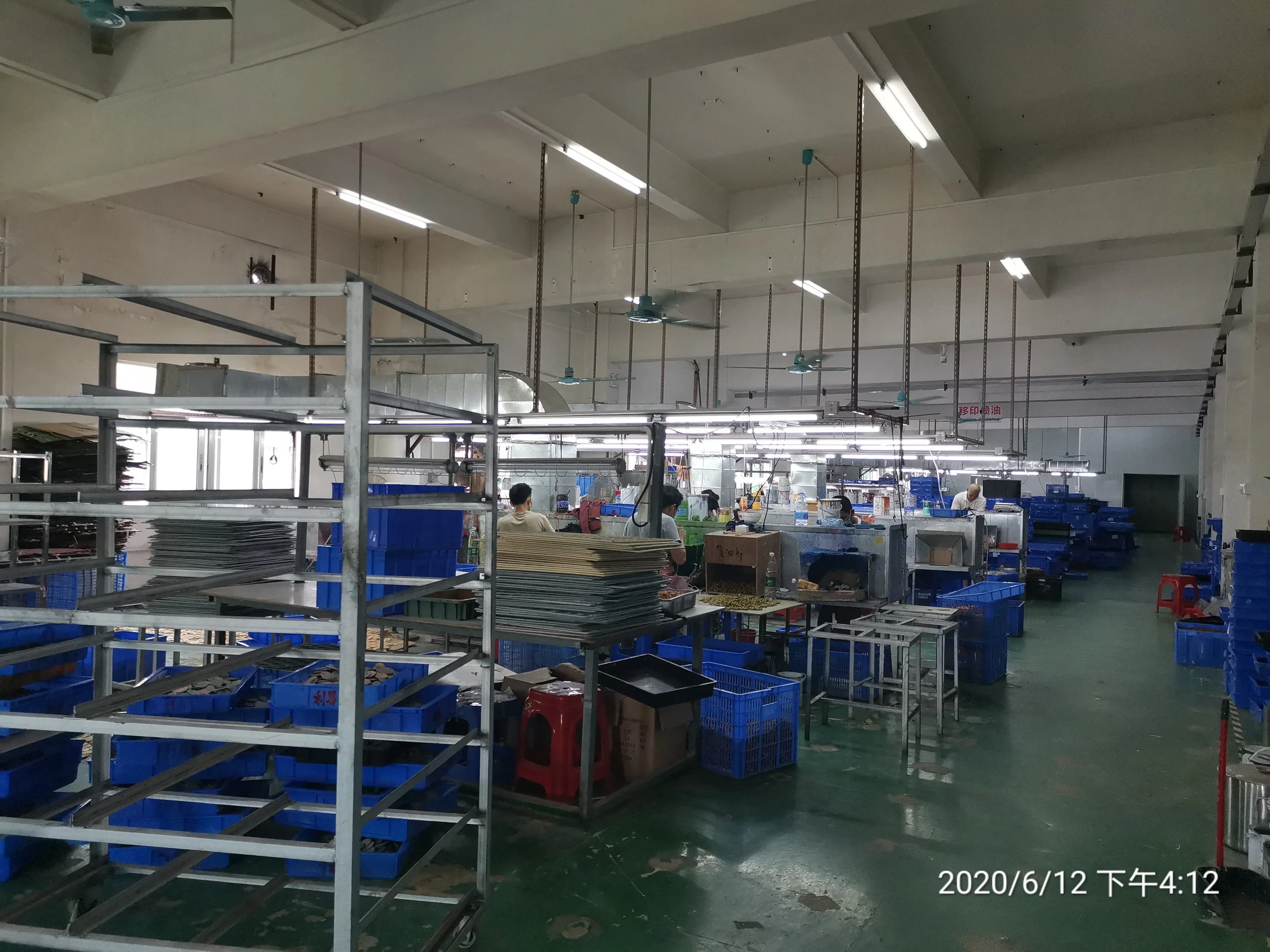 Inside a factory or warehouse with workers and metal shelving units holding papers, blue plastic bins, and other materials. Overhead lighting and ceiling fans are visible.