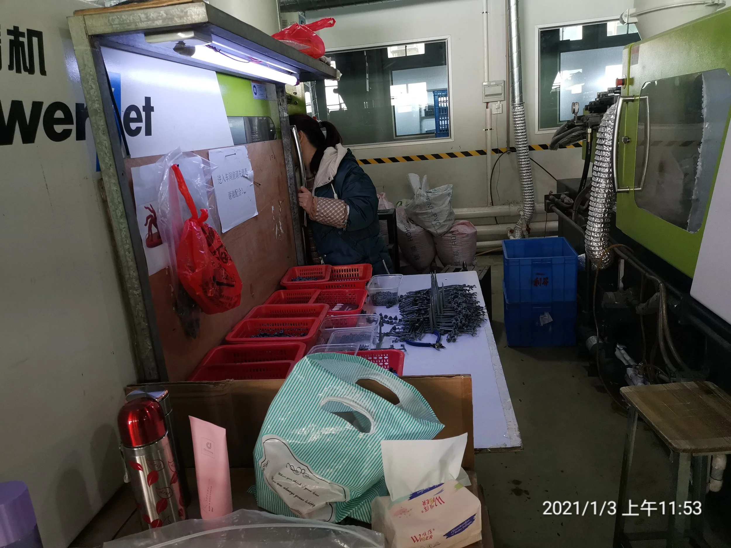 Person working at a machine in an industrial or manufacturing setting, surrounded by various tools, plastic containers, and supplies.