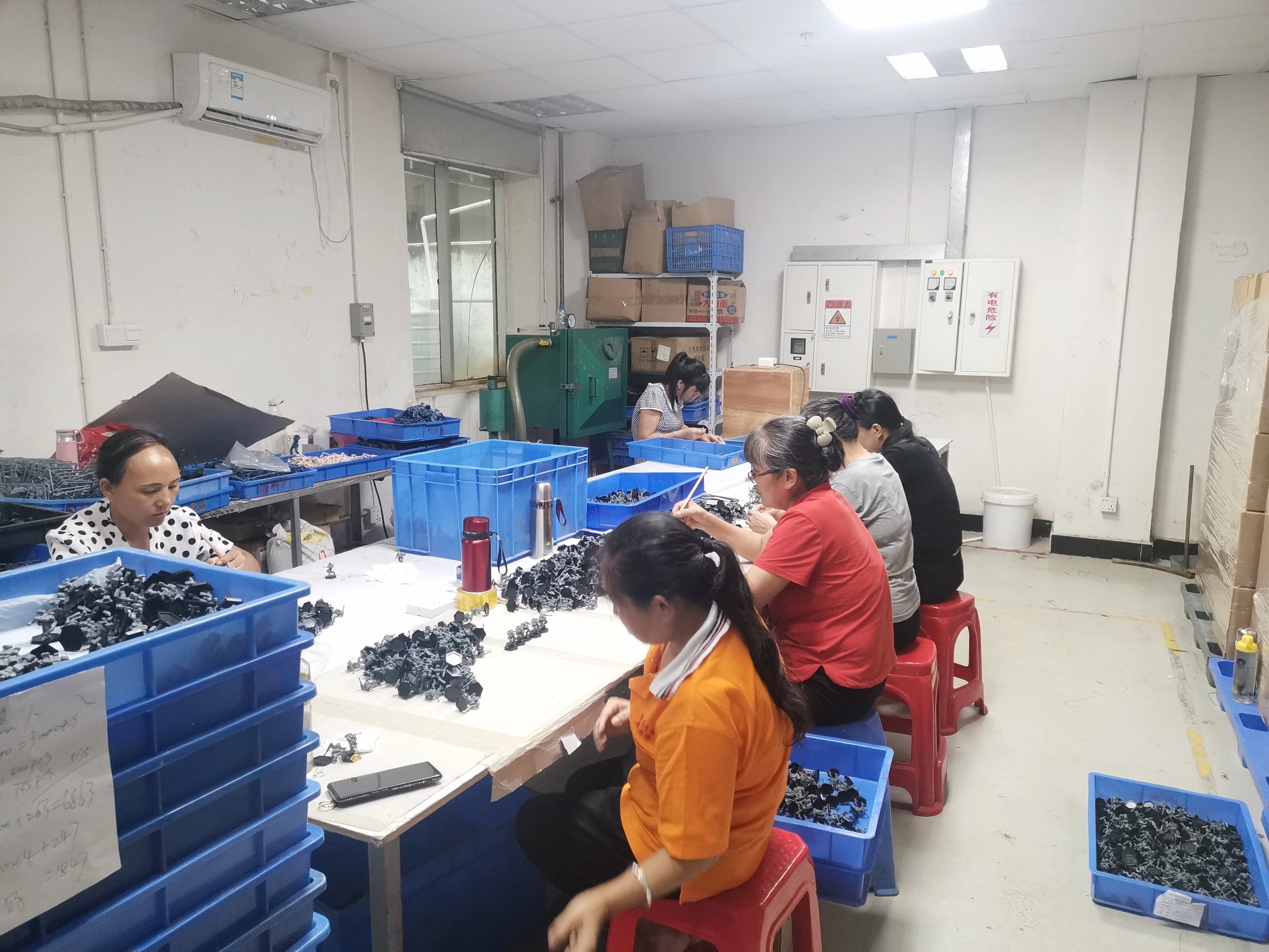 Workers seated on small red stools at a long work table, sorting and assembling small electronic components in a workshop. Large blue plastic bins filled with parts are on the table and nearby.