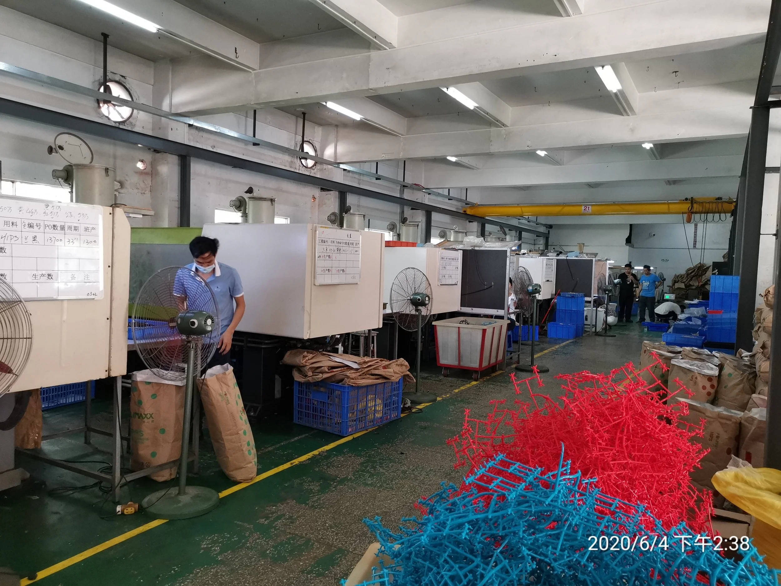 Manufacturing factory with workers operating machines, piles of brown paper bags, and blue and red plastic components on the floor.