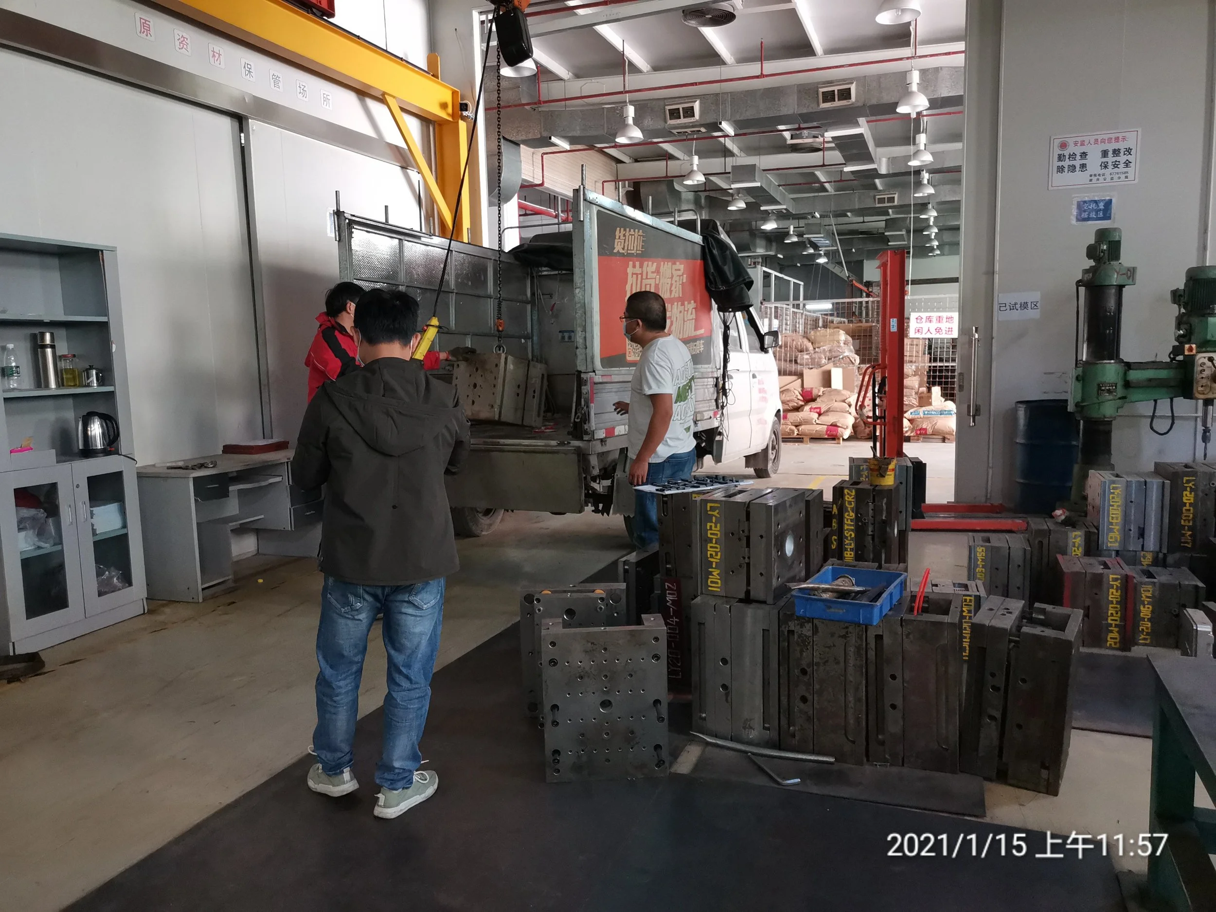 Three people inspecting equipment in a warehouse. One person is wearing a white T-shirt and jeans, while the other two are wearing dark jackets. Various industrial tools and machinery are visible, along with a truck, metal parts, and supplies in the 