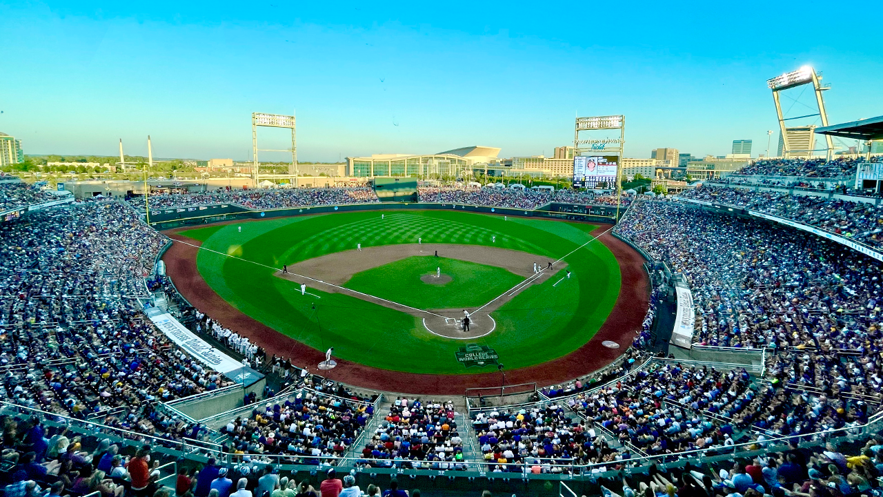 A baseball game in progress at a large stadium filled with spectators, with players on the field and a bright blue sky overhead.