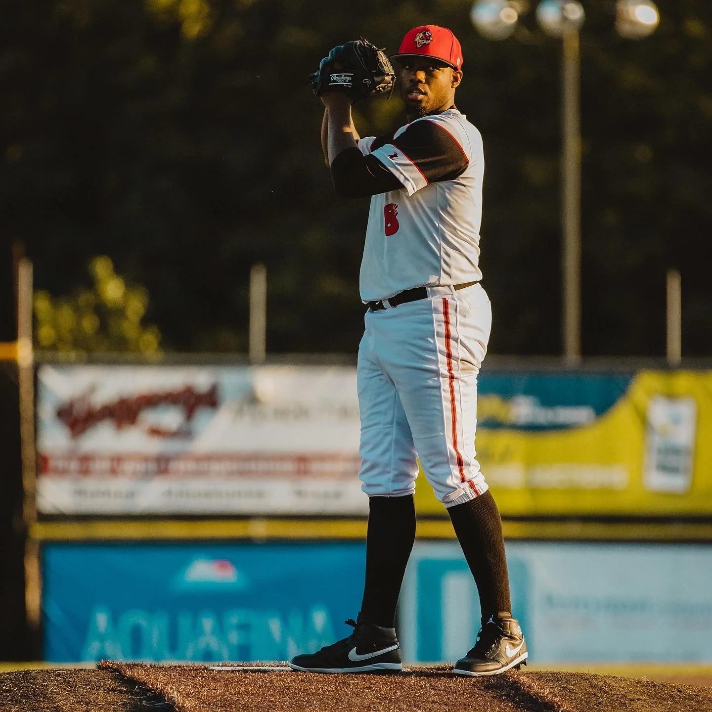A baseball player standing on the pitcher's mound, holding a baseball in his glove, wearing a red cap, white uniform with black and red accents, black socks, and black Nike shoes.