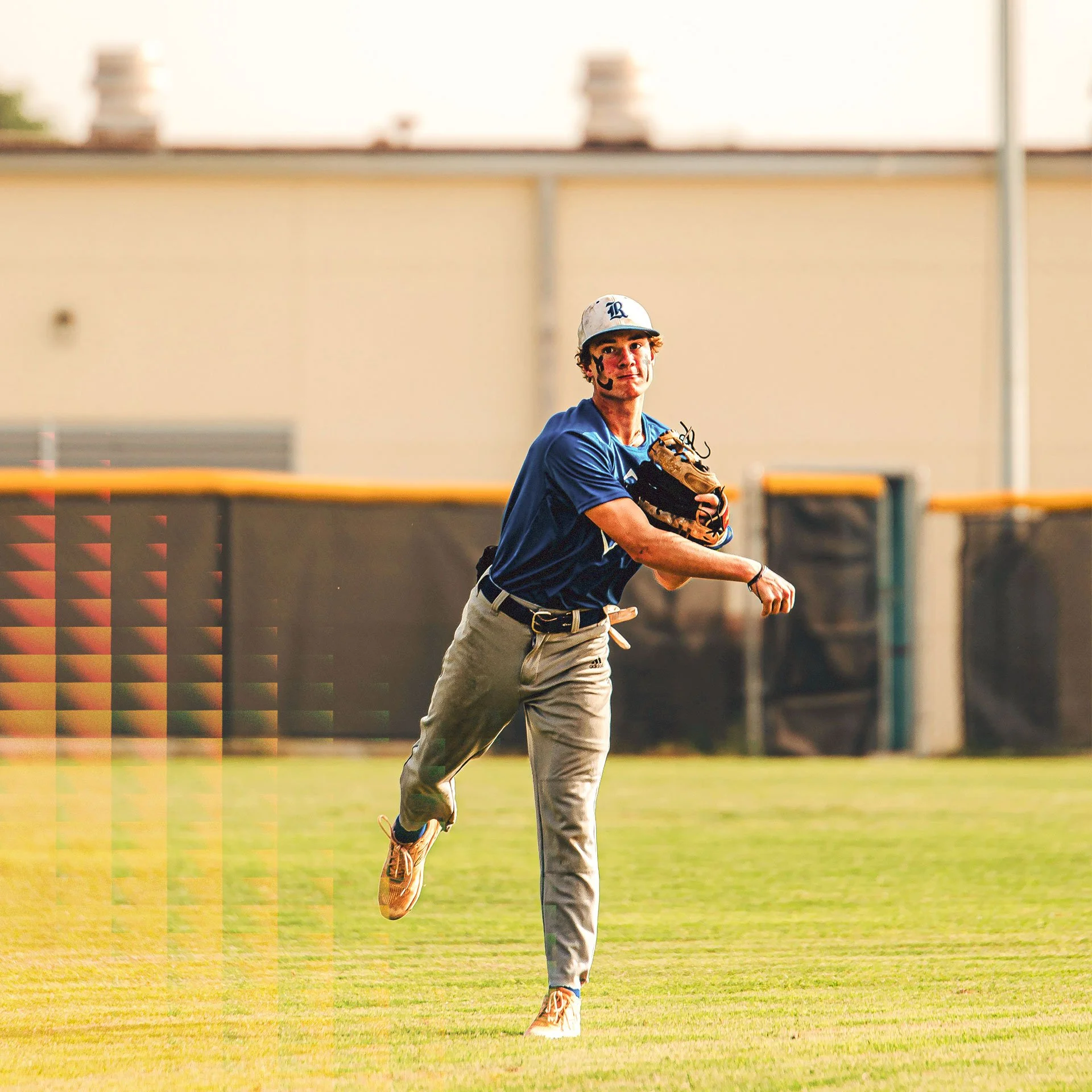 A young male baseball player in a blue shirt and khaki pants running on the grassy field while holding a baseball glove, with a stadium fence and building in the background.