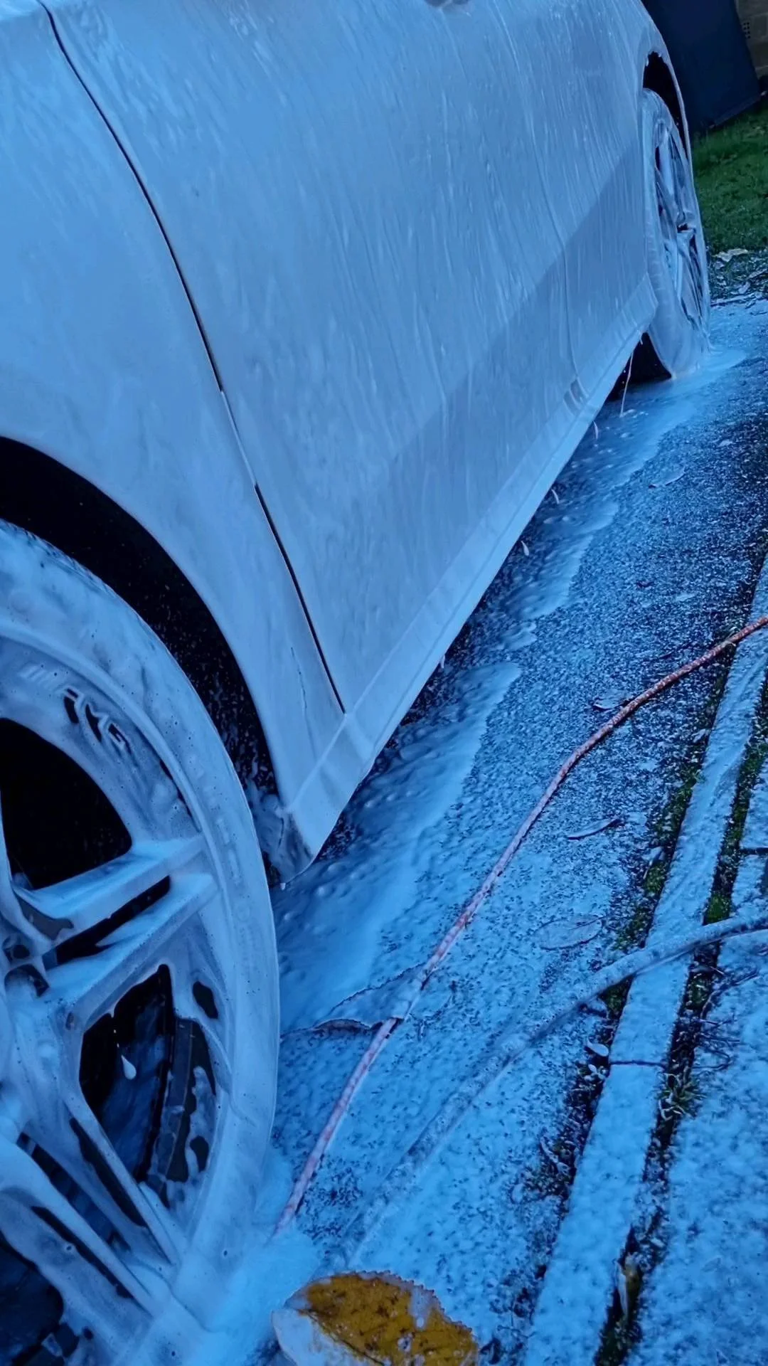 A white car is being washed with soap suds and water, lying on a wet surface with foam and a hose nearby.