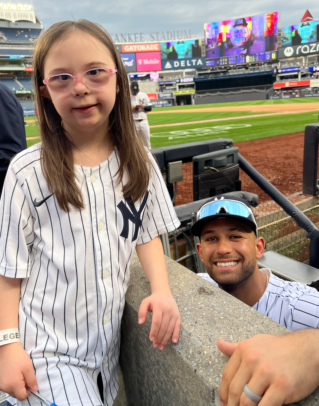 A young girl and a man at a baseball stadium, both wearing New York Yankees jerseys. The girl is standing near the barrier, and the man is crouching beside her, smiling for the photo. The stadium is visible in the background, with the field and elect