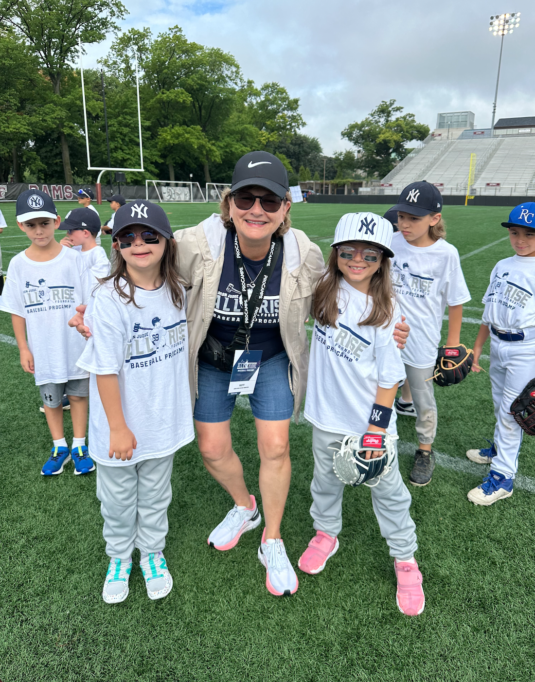A group of young children and an adult woman on a baseball field, wearing shooting shirts and caps, some with sunglasses, posing for a photo.