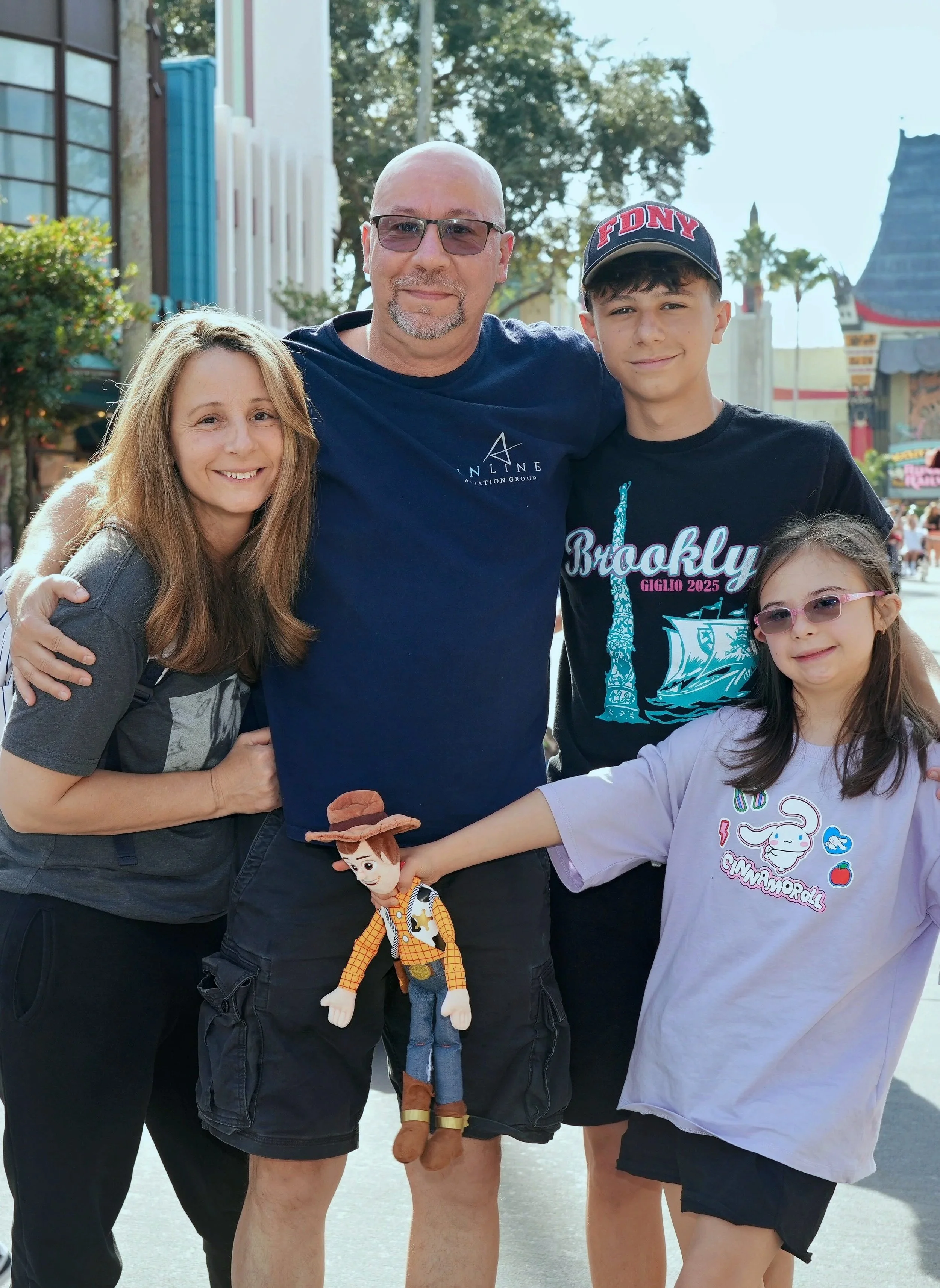 A family of four posing together at an amusement park, with a toy cowboy doll held by the man in the center.