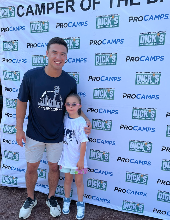 A man and a young girl are standing in front of a Dick's Sporting Goods backdrop. The man is wearing a navy t-shirt and white shorts, and the girl is wearing a white t-shirt and colorful shorts. They are smiling and posing for the photo.