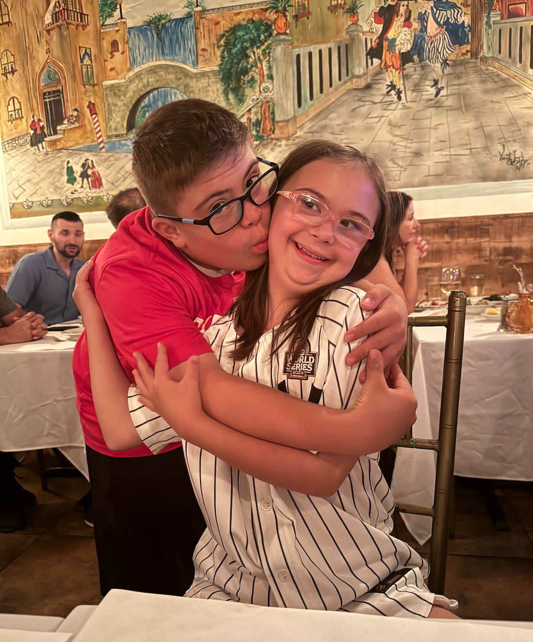 A boy and girl hugging at a restaurant, with the boy kissing the girl's cheek. The girl is smiling and wearing glasses, and both children have glasses. The background shows other diners and a mural painting on the wall.