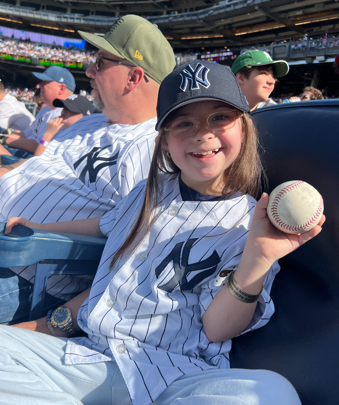 Young girl with glasses wearing a New York Yankees cap holding a baseball at a baseball game.